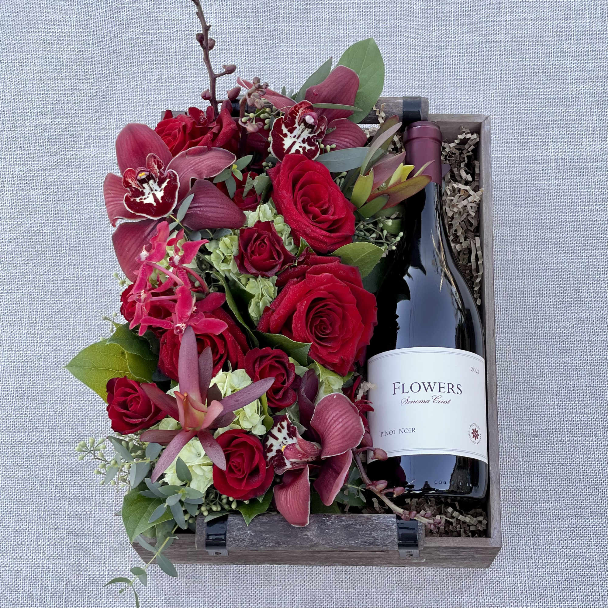 Red roses and orchids arranged beside a bottle of pinot noir in a wooden gift box.