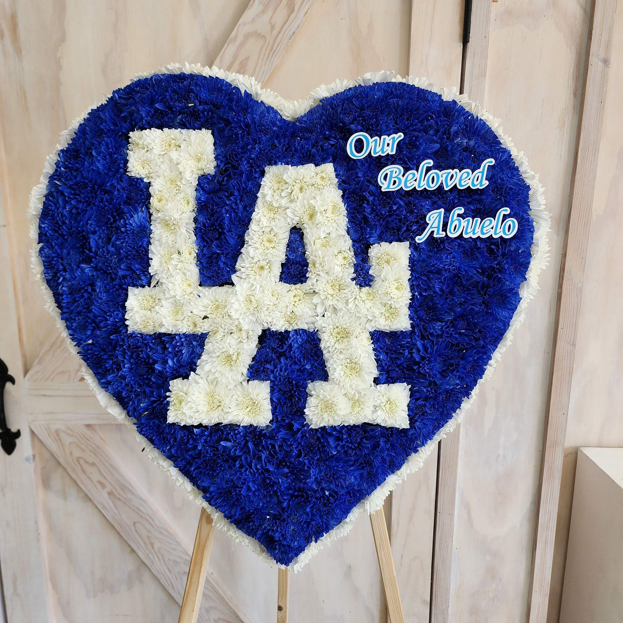 Heart-shaped floral tribute with blue and white flowers reading "LA" and "Our Beloved Abuelo"