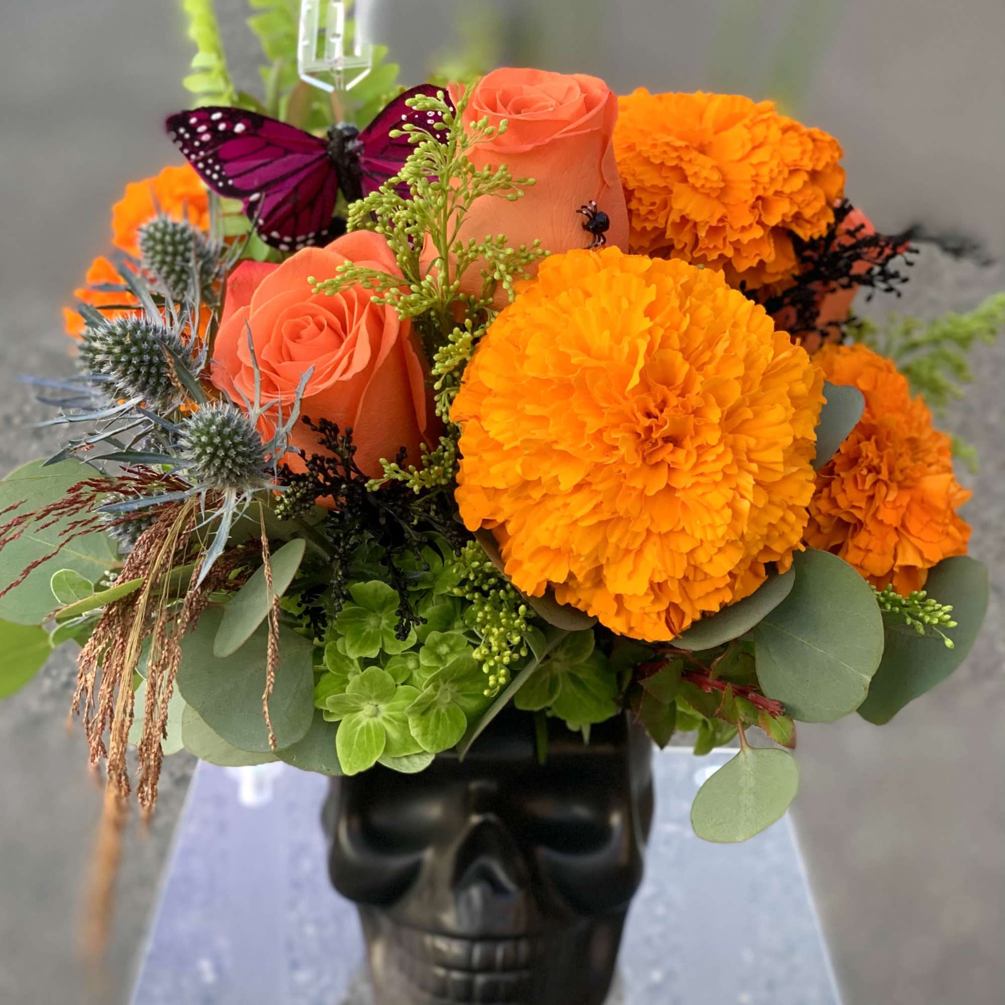 Orange roses and marigolds arranged in a black skull vase with a purple butterfly
