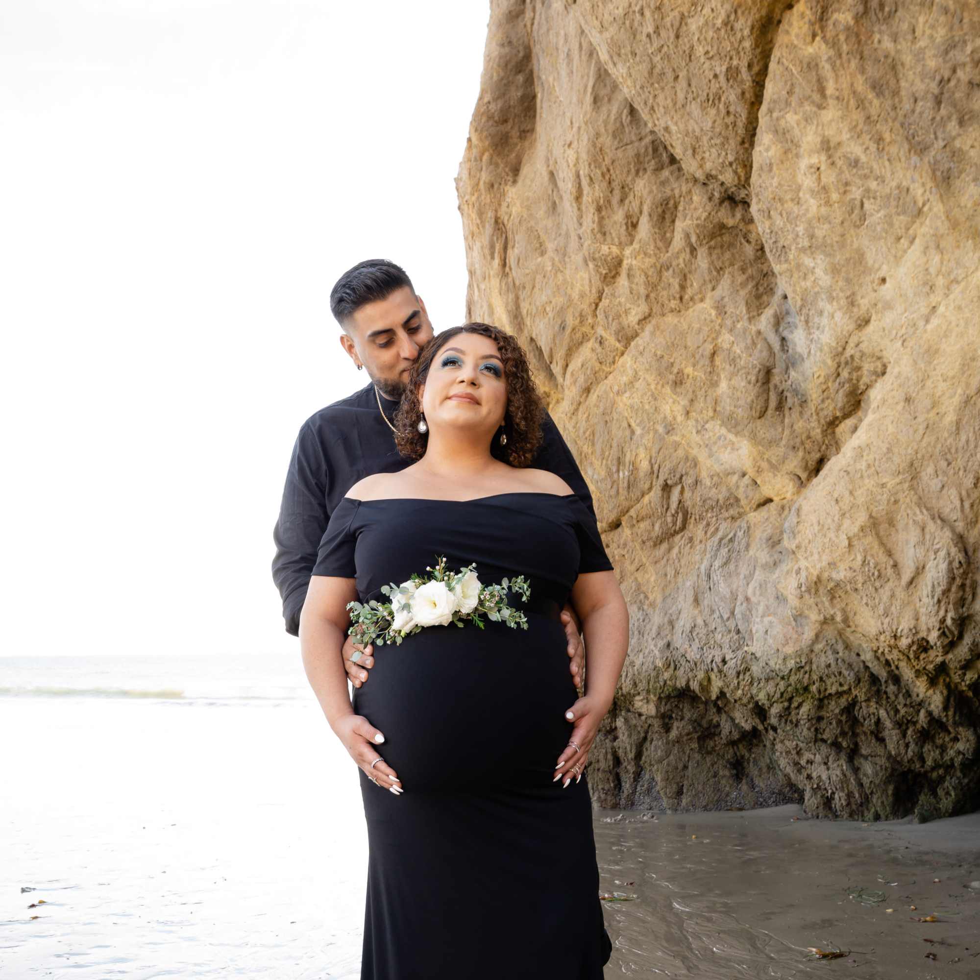 Pregnant woman in a black dress with a white floral belt, standing by a rock wall