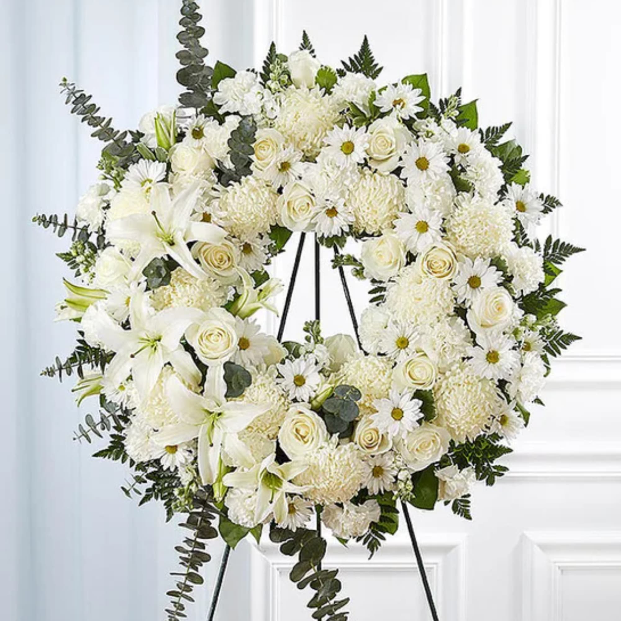White floral wreath on a black stand with lilies, roses, and daisies