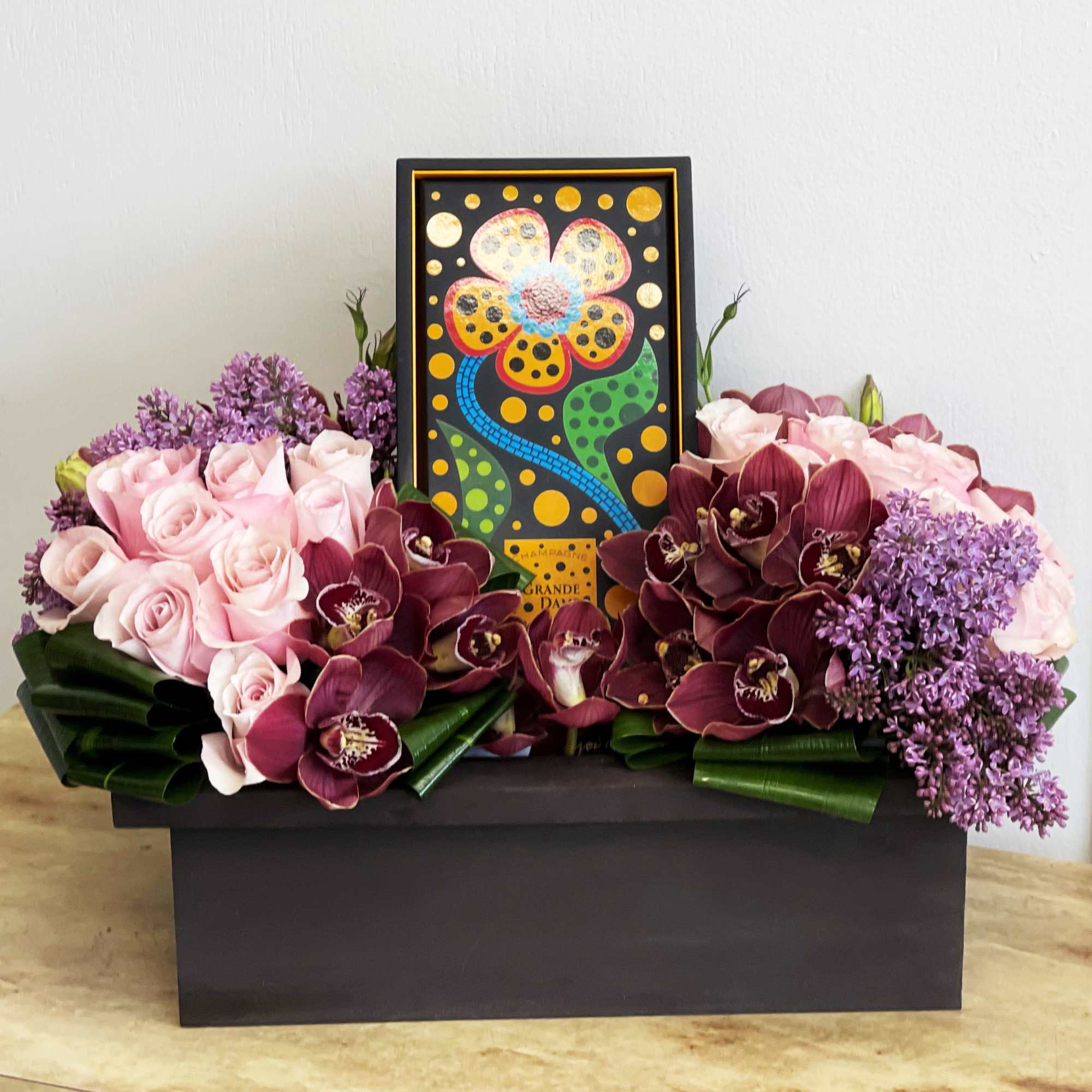 Pink roses and burgundy orchids arranged around a framed floral artwork in a black box