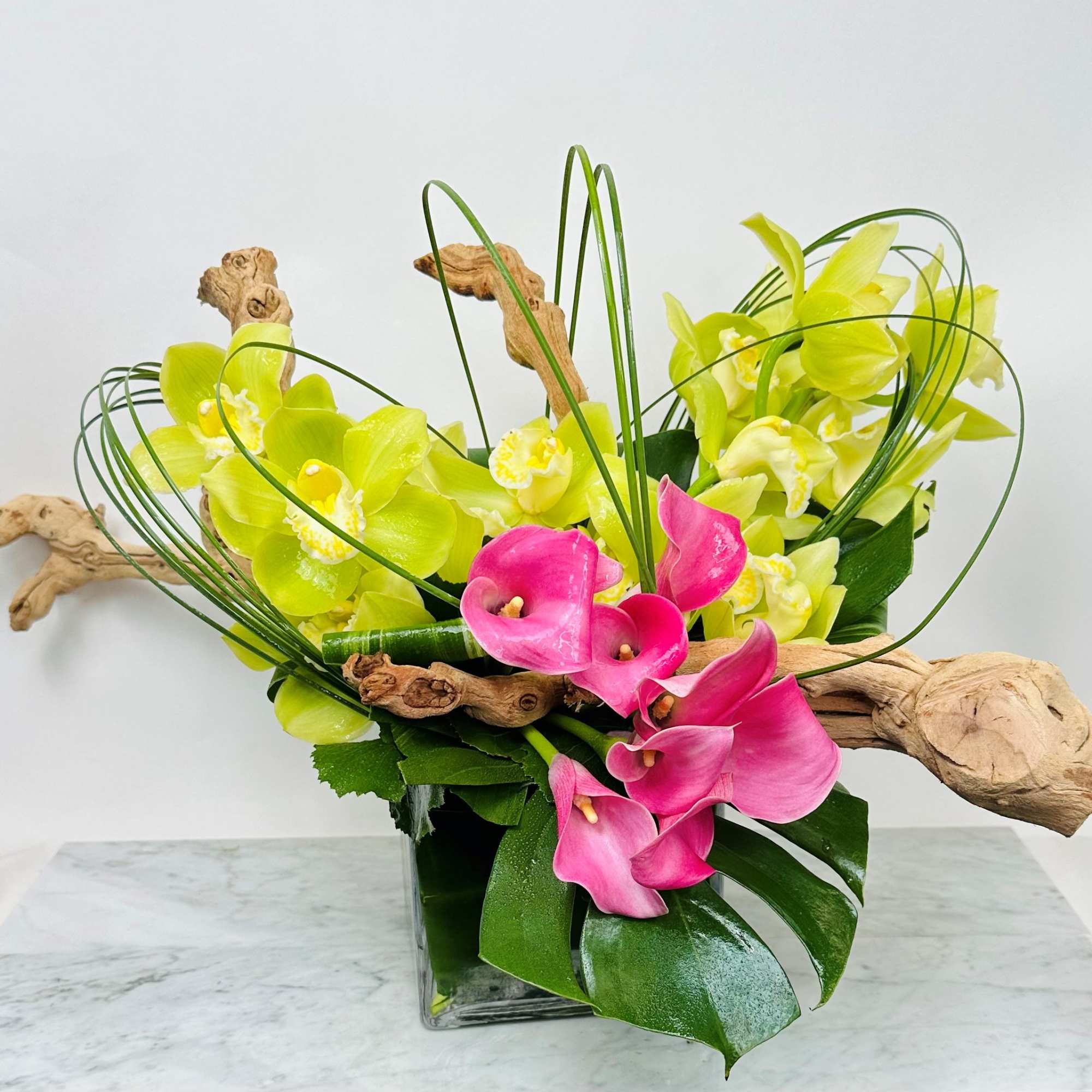 Pink and yellow orchids arranged in a square glass vase with curly branches