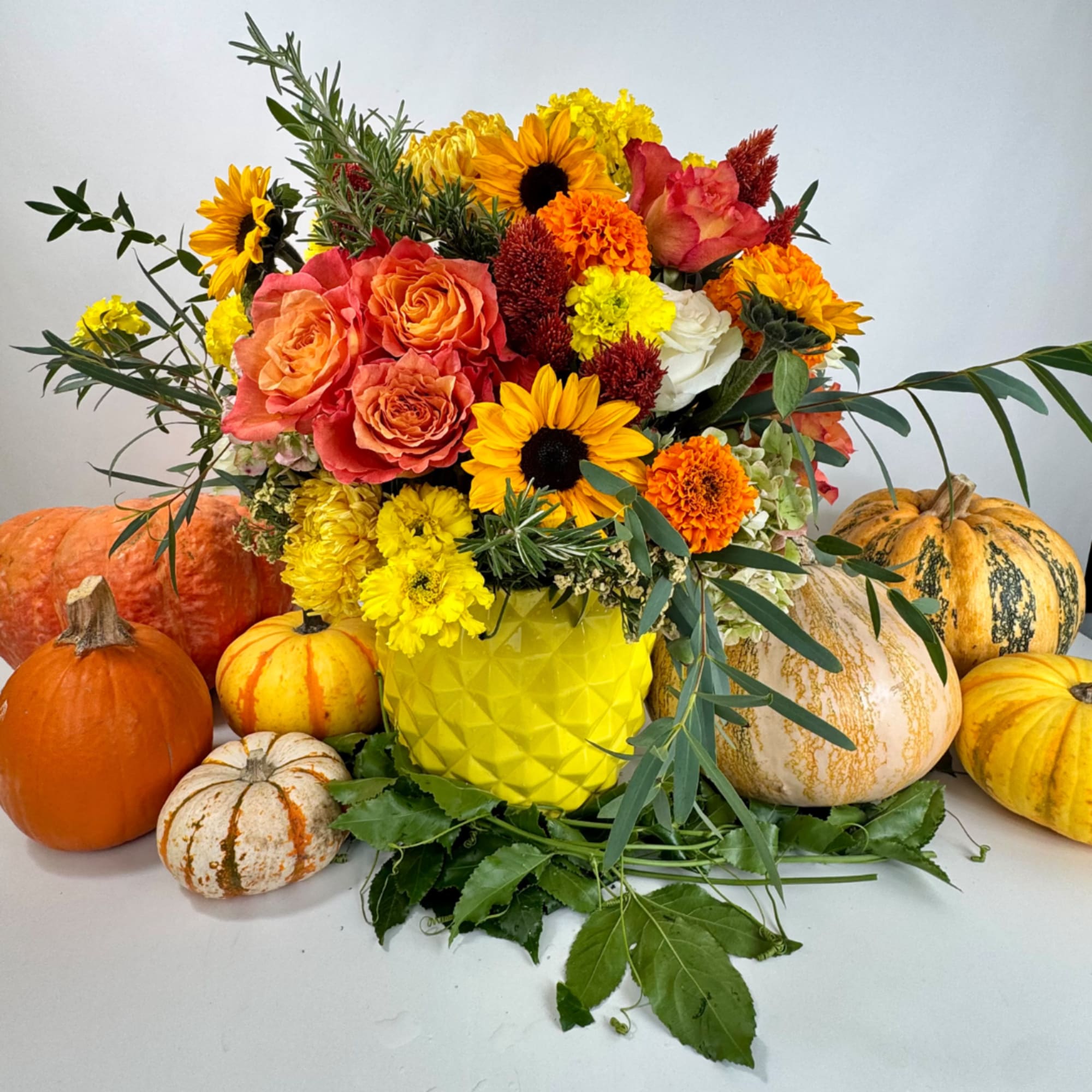 Autumn bouquet with roses, sunflowers, and mums in a yellow vase surrounded by pumpkins