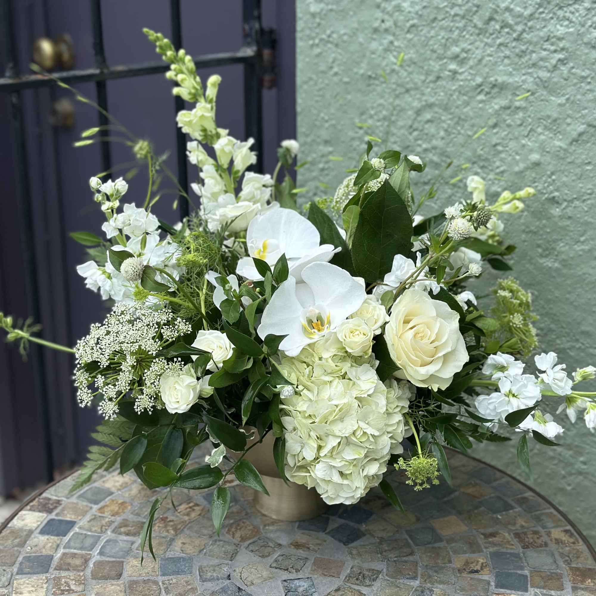 Low white arrangement with roses, orchids, hydrangeas, and snapdragons in a gold vase