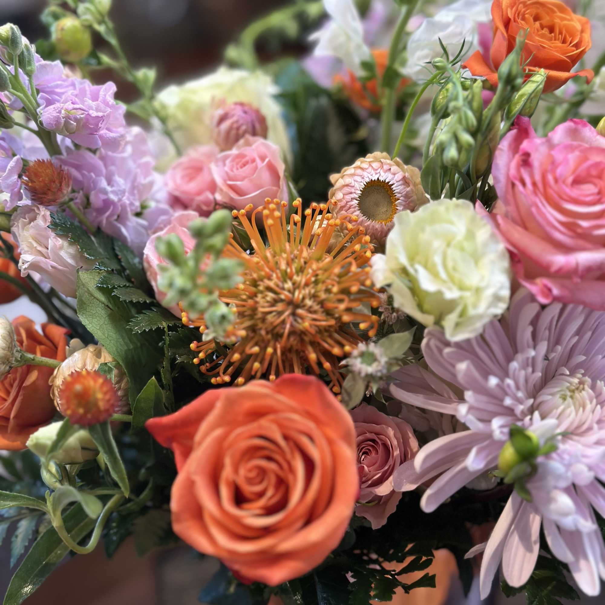 Colorful arrangement with orange roses, pincushion protea, pink roses, and soft lavender blooms in a vase