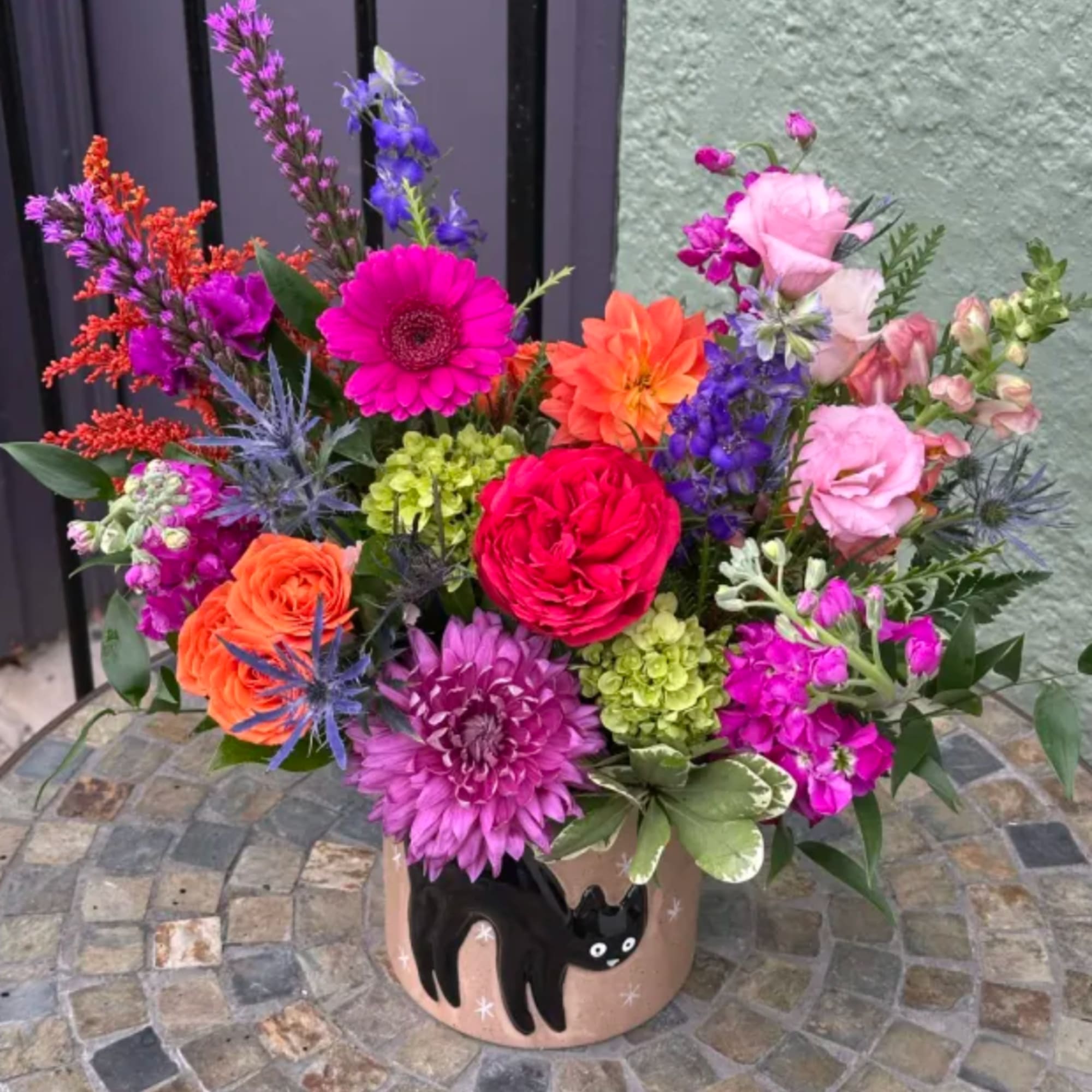 Bright mixed bouquet in a cat-decorated pot with pink, orange, purple, and green blooms.