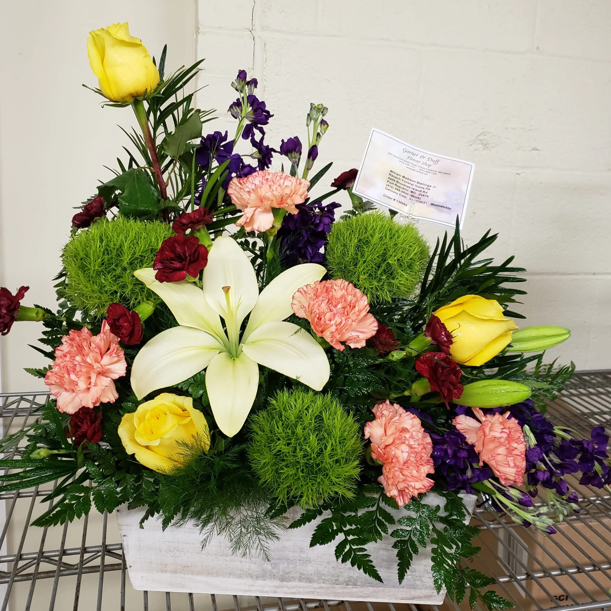 Mixed floral arrangement with yellow roses, white lilies, and pink carnations in a white container