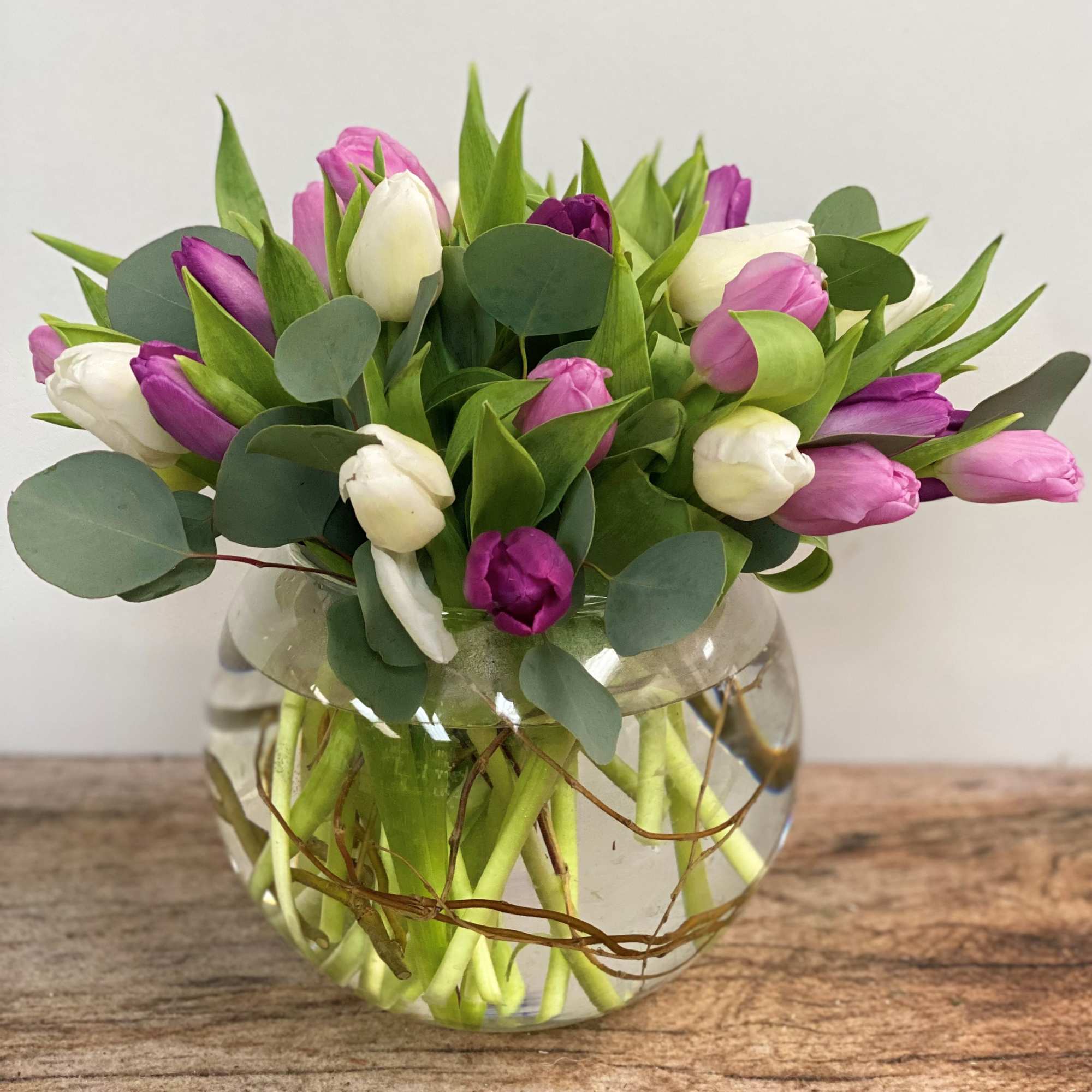 Round glass vase of white and purple tulips with eucalyptus accents on a wooden surface.
