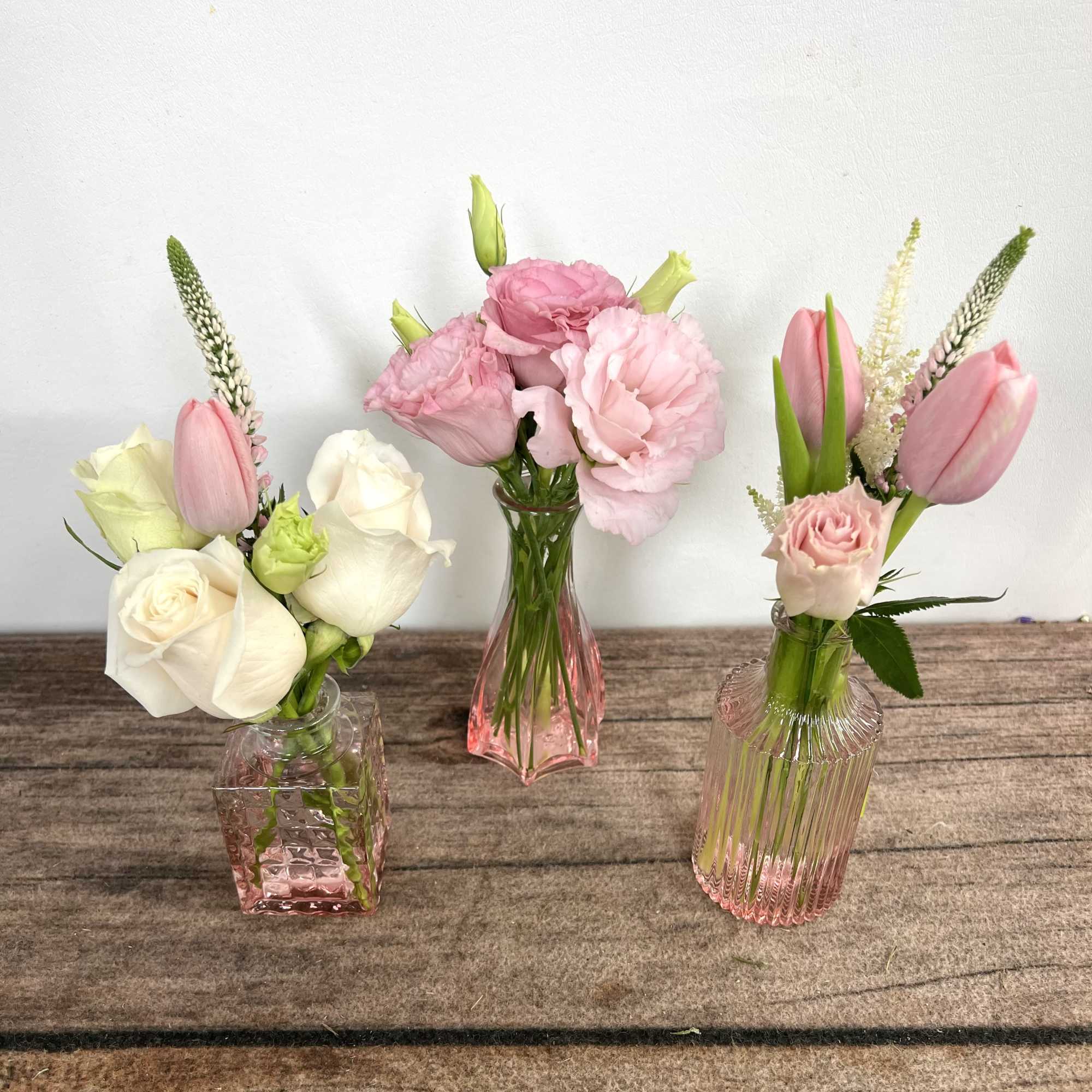 Three small pink glass bud vases with white roses, pink tulips, and pink lisianthus on a wooden surface.