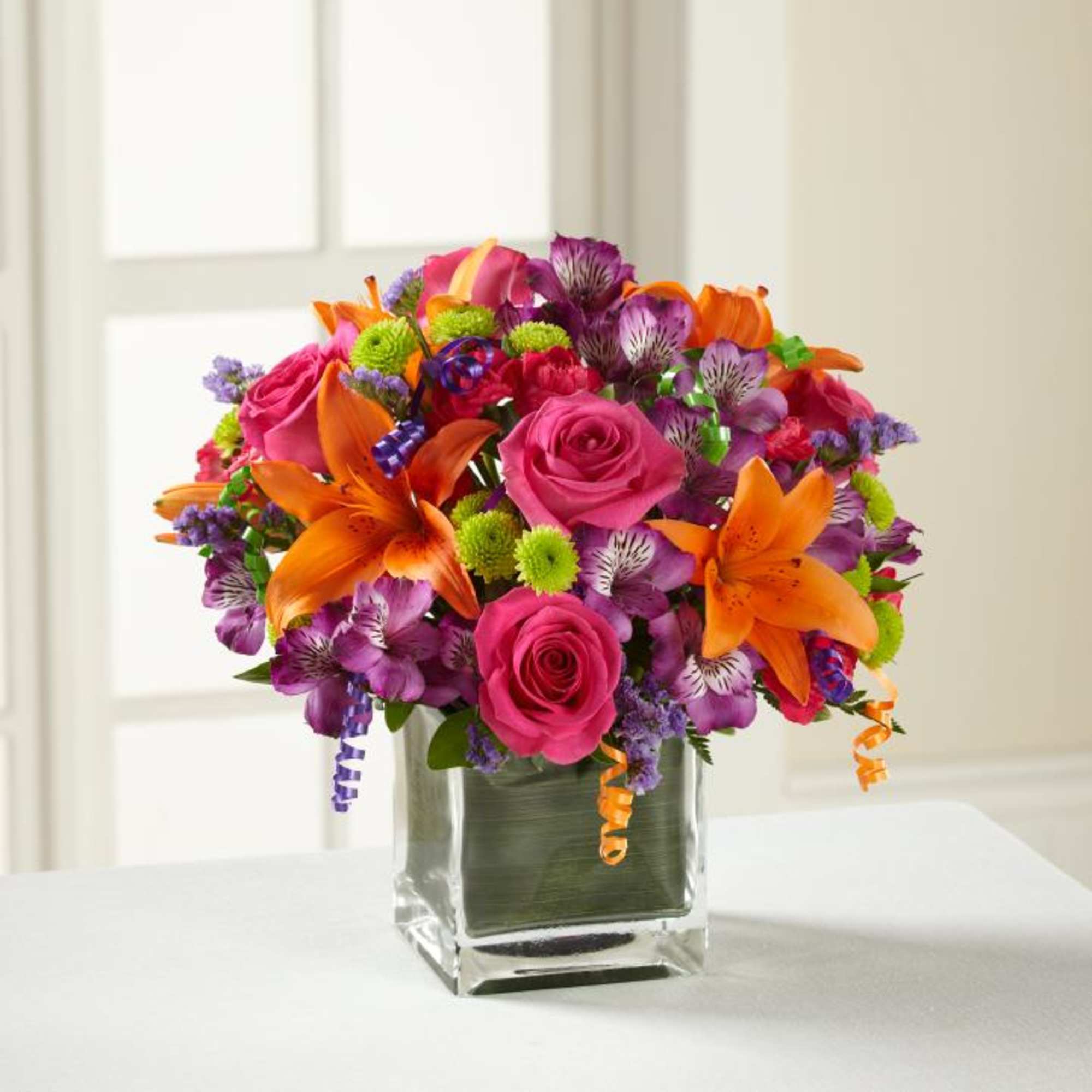 Bright mixed bouquet of pink roses, orange lilies, and purple flowers in a glass vase