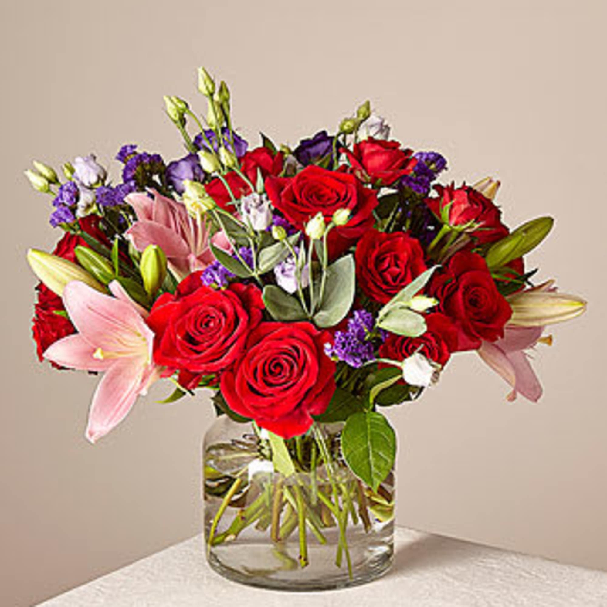 Bouquet of red roses, pink lilies, and purple accents in a glass vase