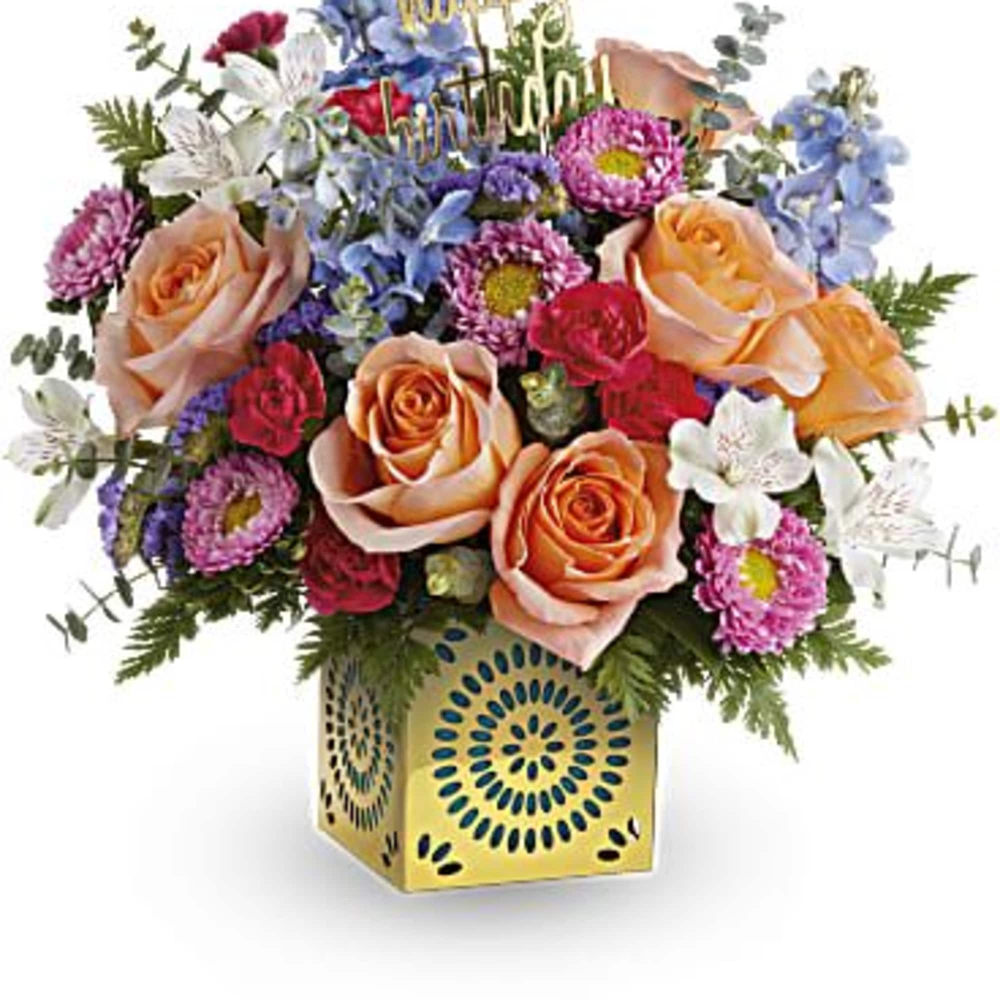 Colorful bouquet of peach roses and mixed blooms in a yellow cube vase with a happy birthday pick.