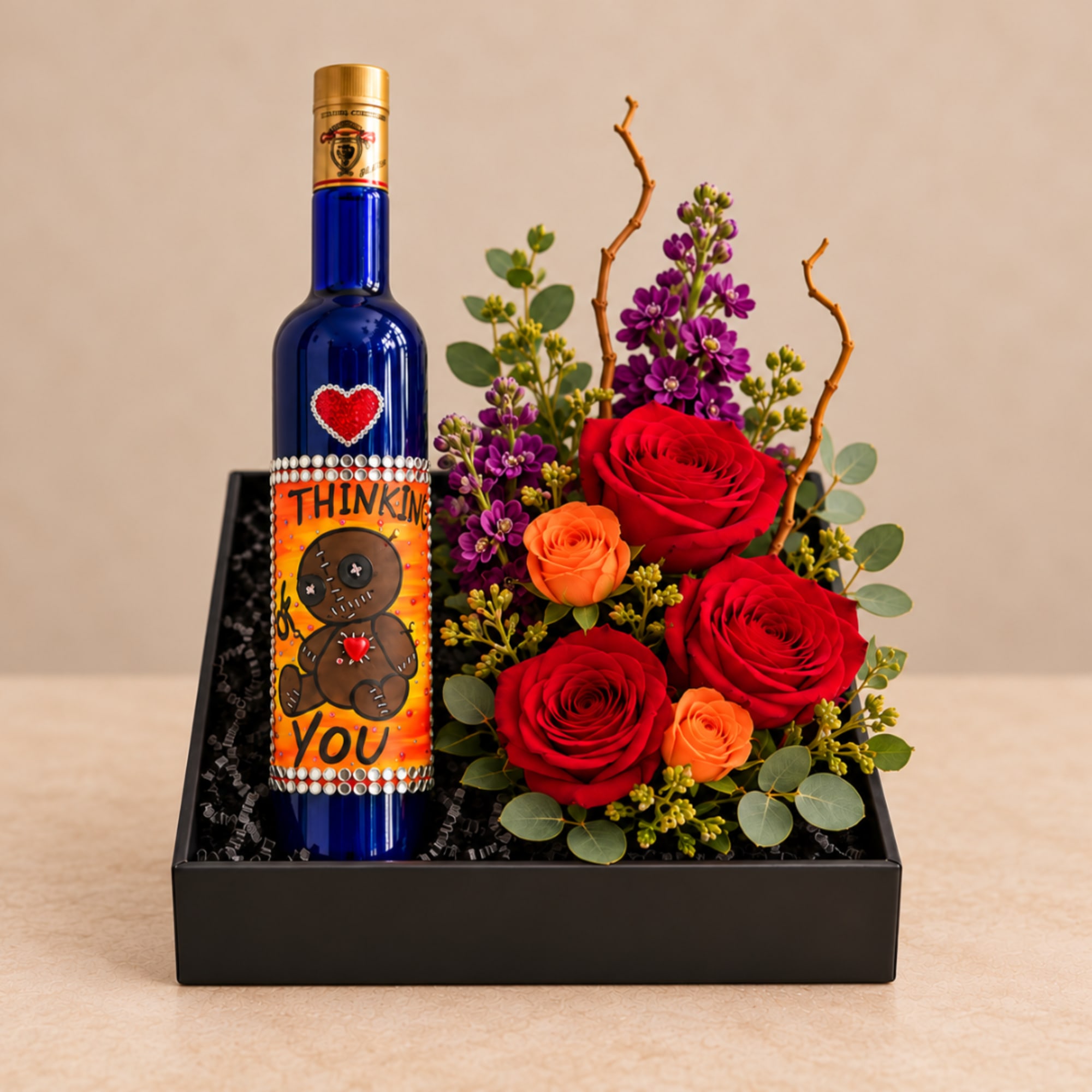 Roses and purple flowers arranged in a black gift box beside a decorated bottle