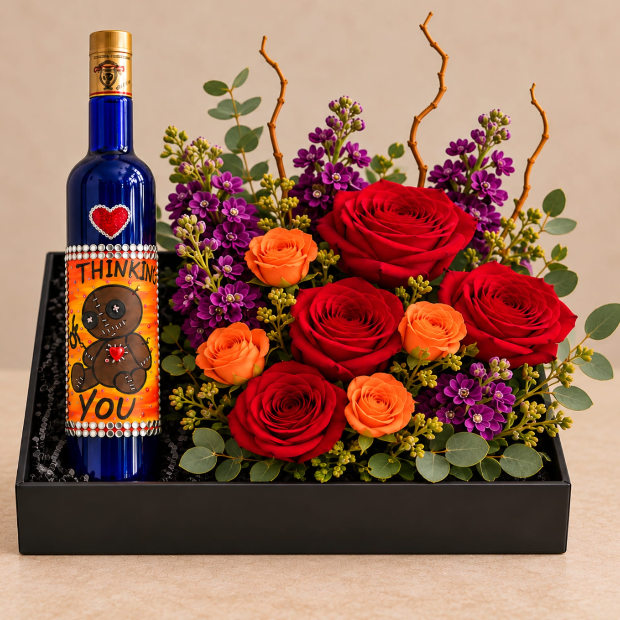 Red and orange roses with purple flowers in a black gift box beside a decorated bottle