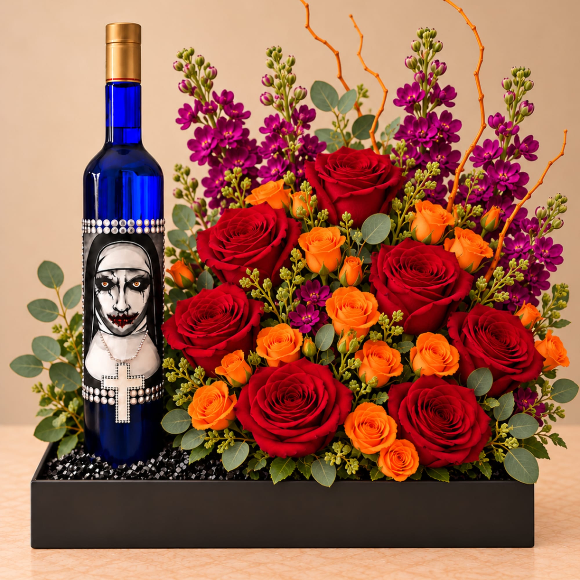Red and orange rose arrangement with purple flowers in a black box beside a decorated blue bottle