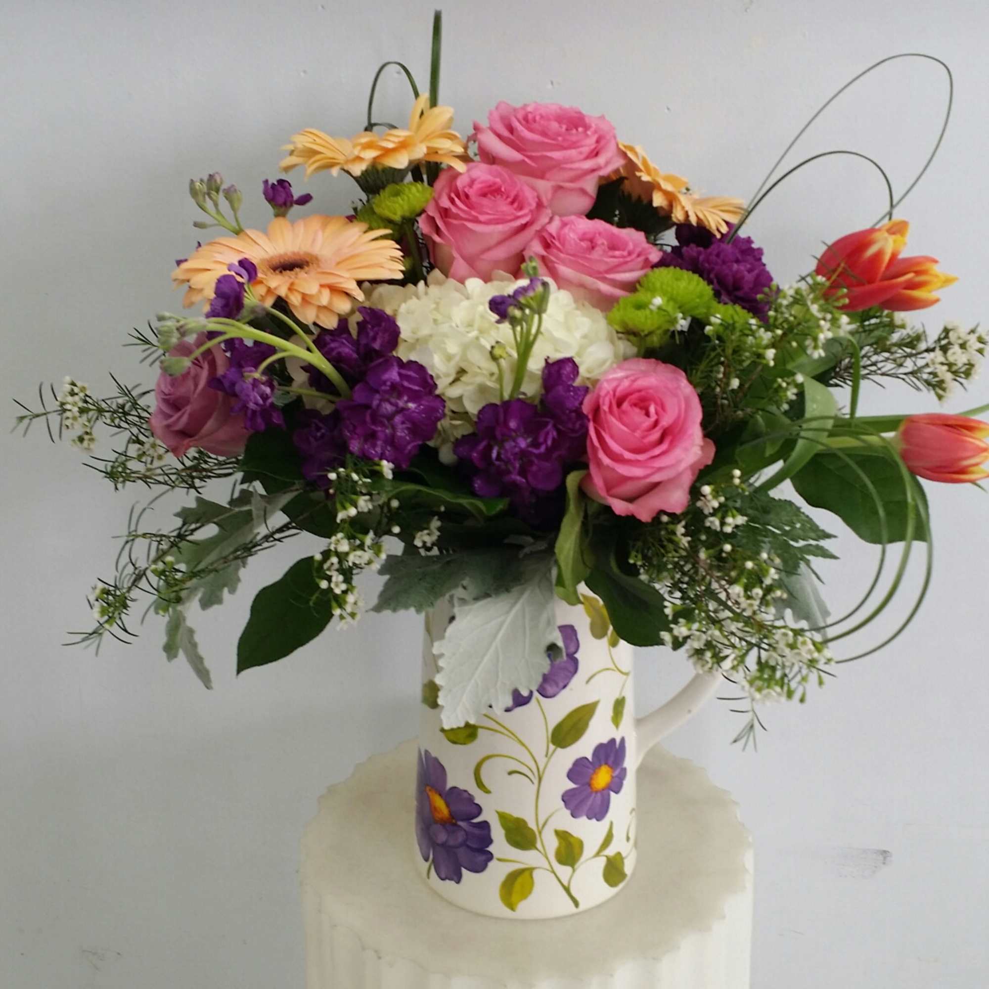 Mixed bouquet of pink roses, purple flowers, and orange daisies in a floral pitcher