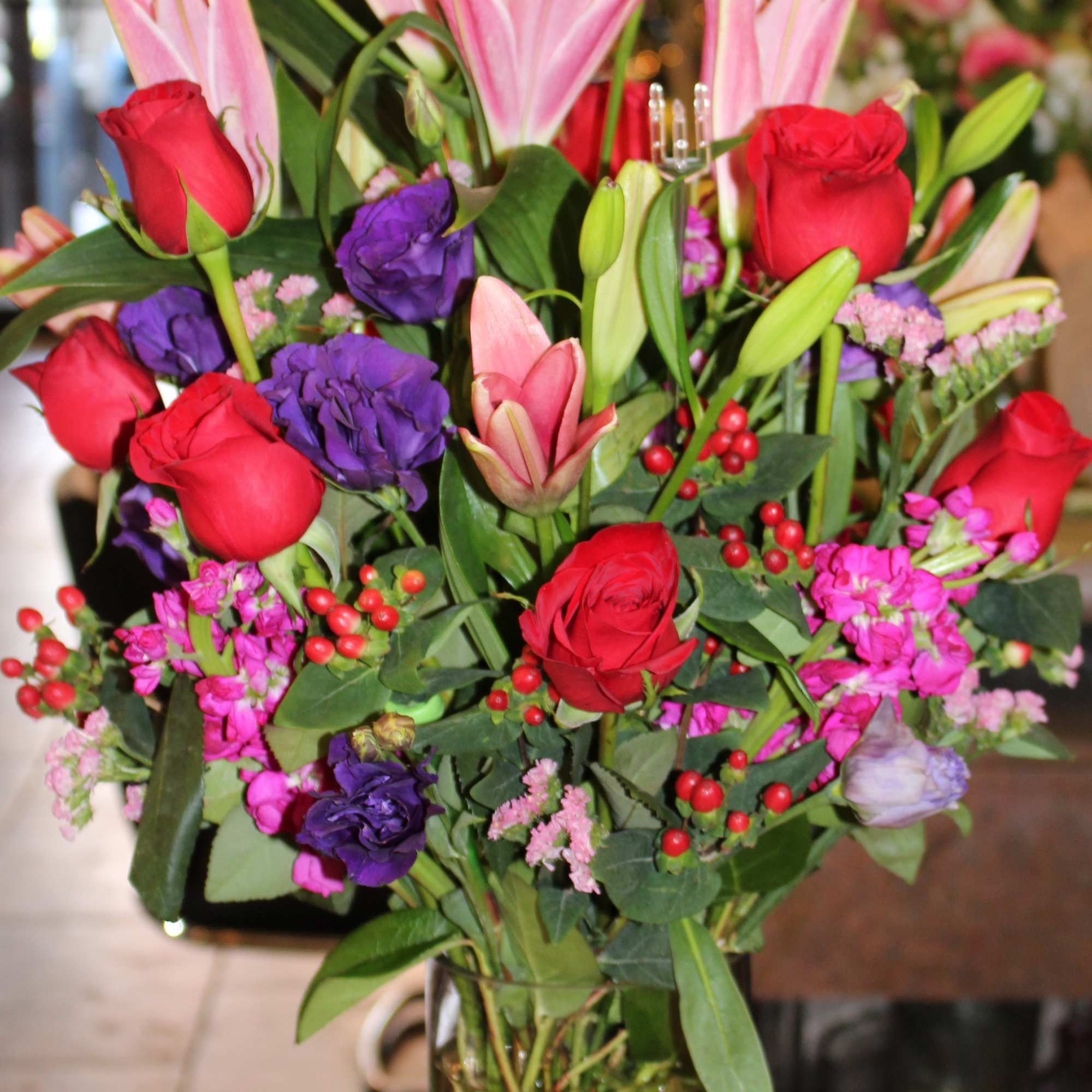 Mixed bouquet of pink lilies, red roses, and purple flowers in a glass vase