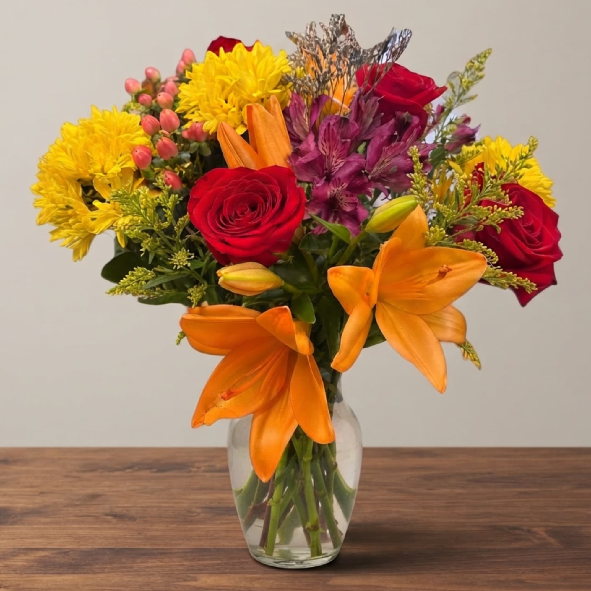 Mixed bouquet of red roses, orange lilies, and yellow mums in a clear vase