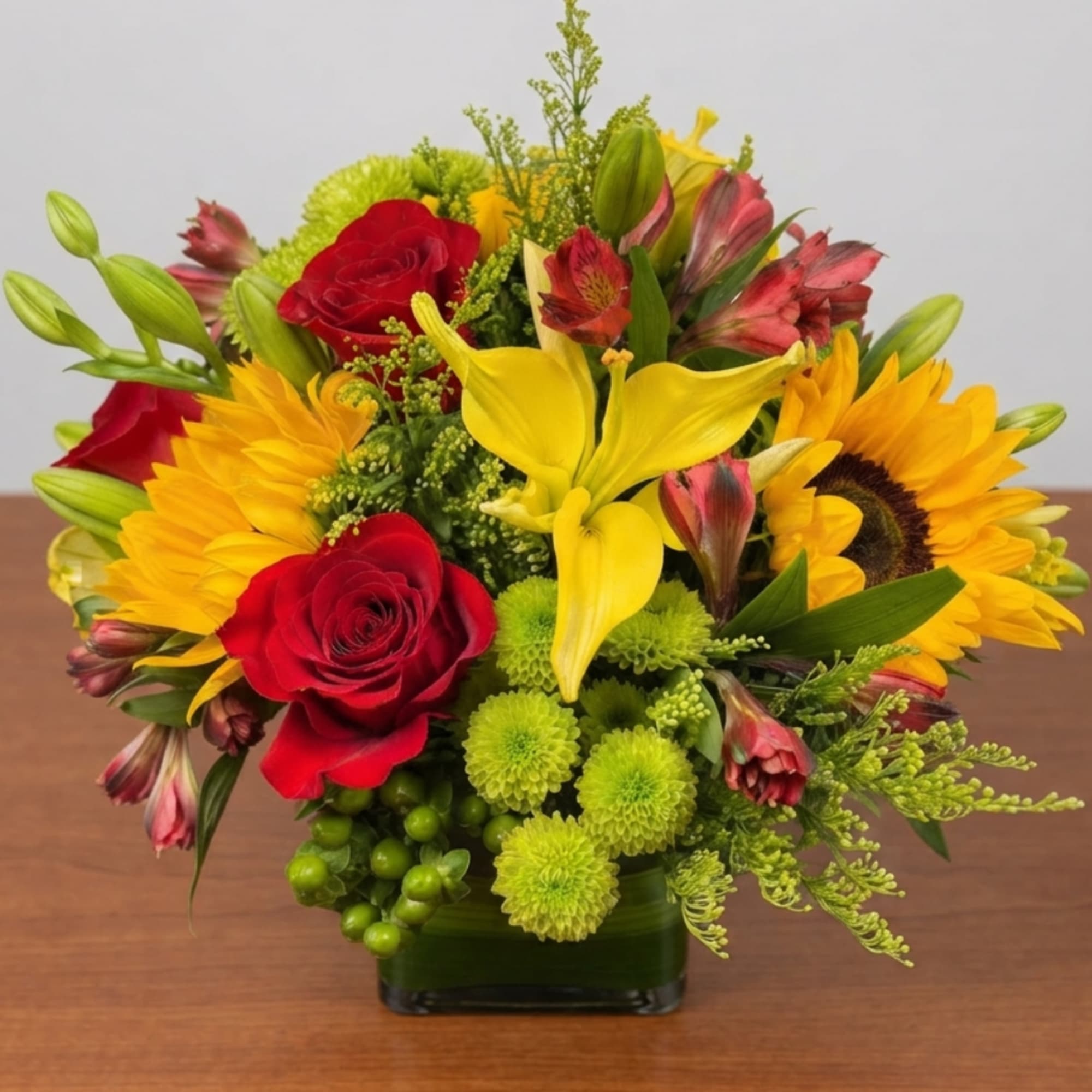 Mixed bouquet with red roses, yellow lilies, and sunflowers in a square vase