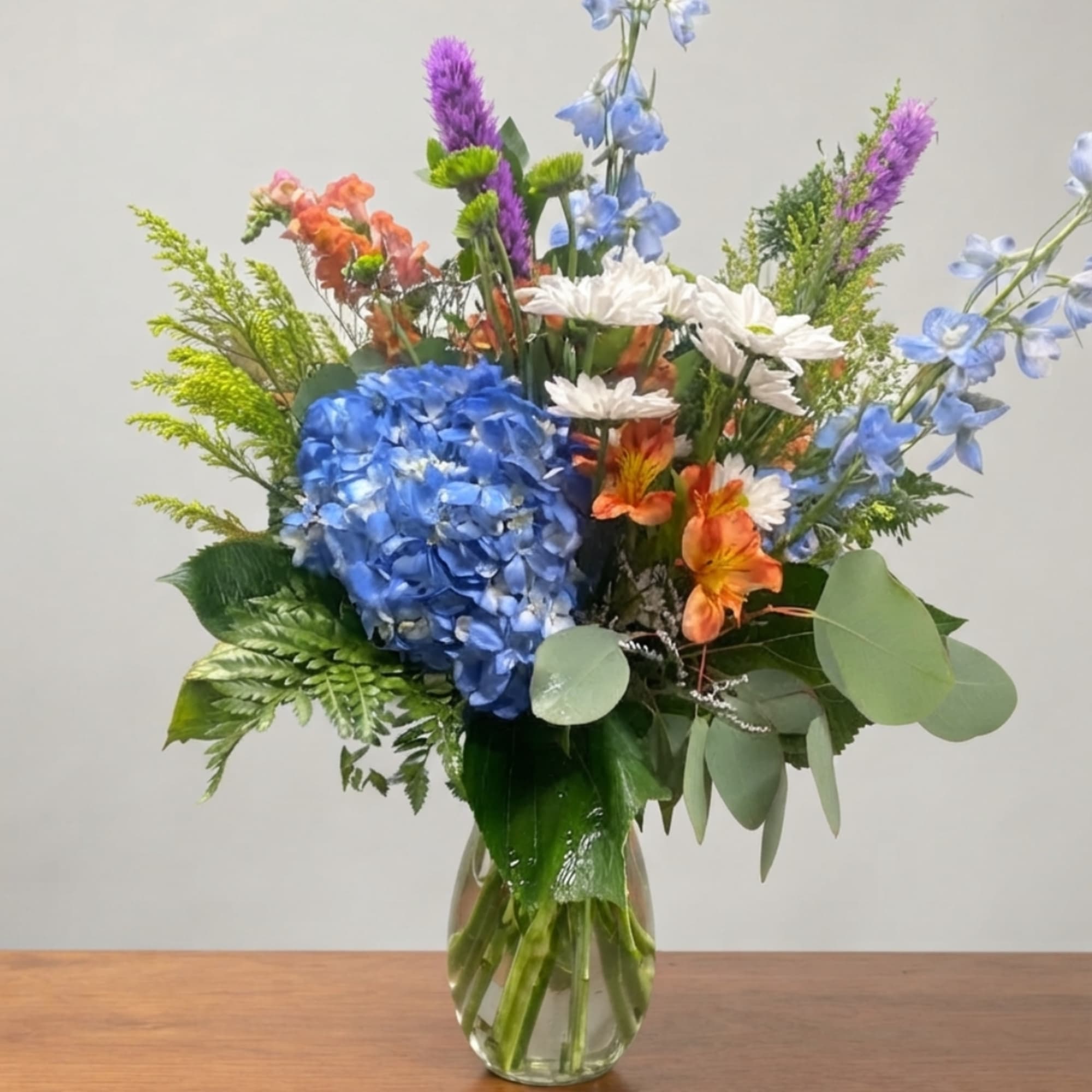 Mixed bouquet with blue hydrangea, white daisies, and orange flowers in a glass vase
