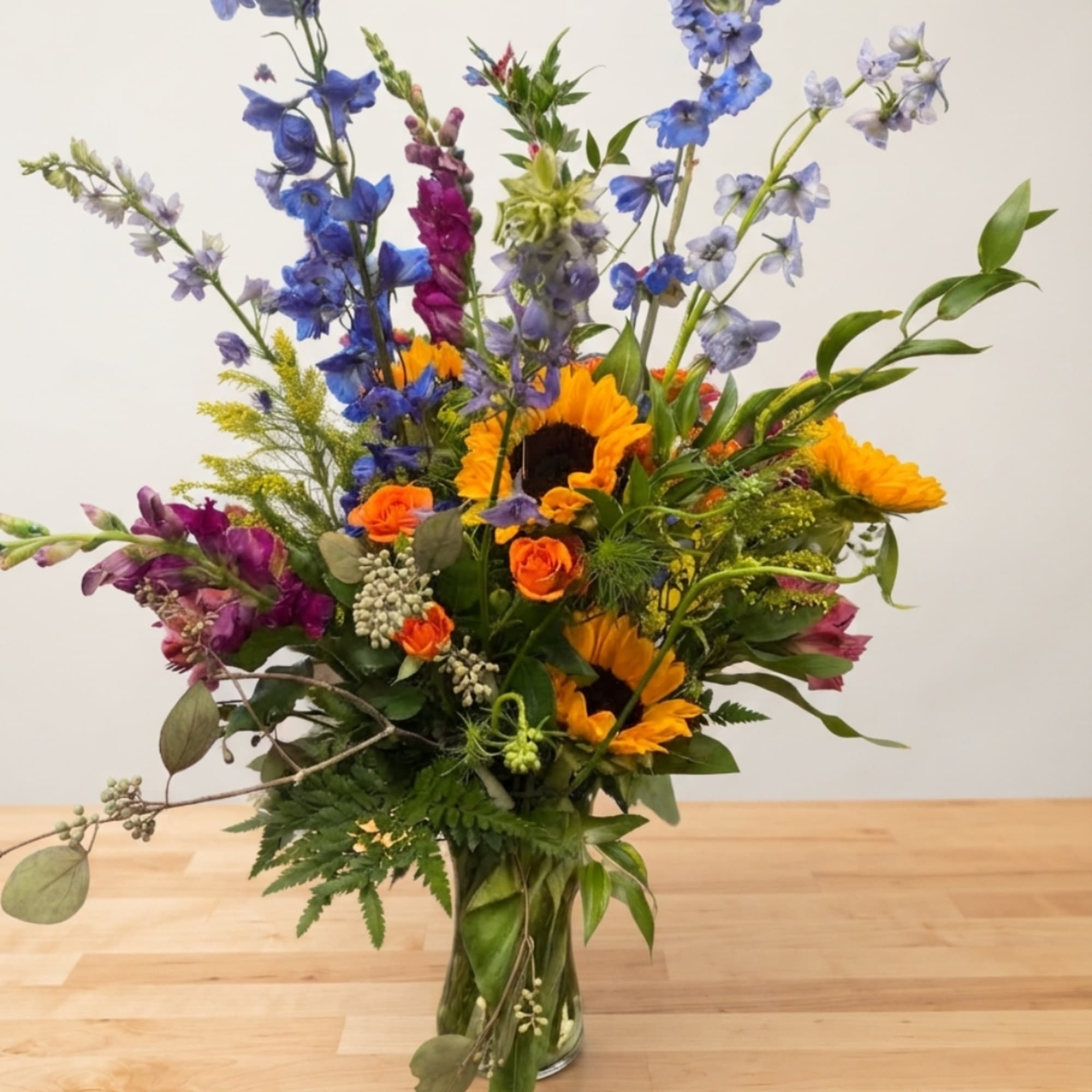 Tall mixed bouquet in a clear glass vase with sunflowers, blue delphinium, and orange roses