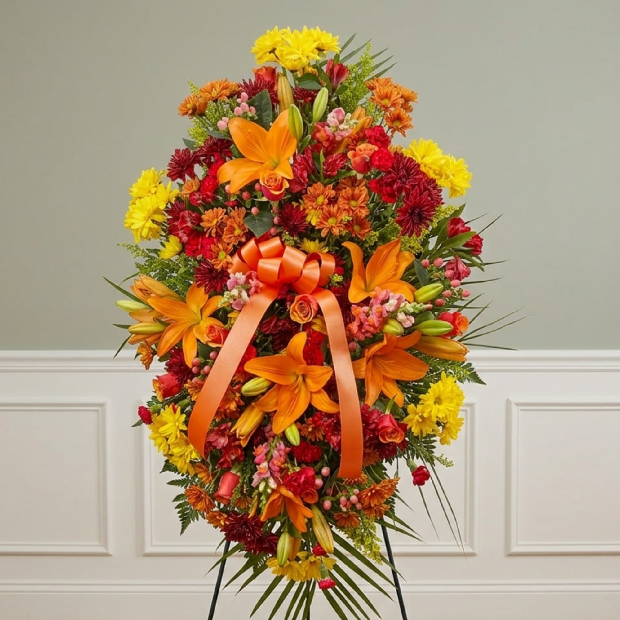 Tall floral standing spray with orange lilies, yellow and red mums, and an orange ribbon