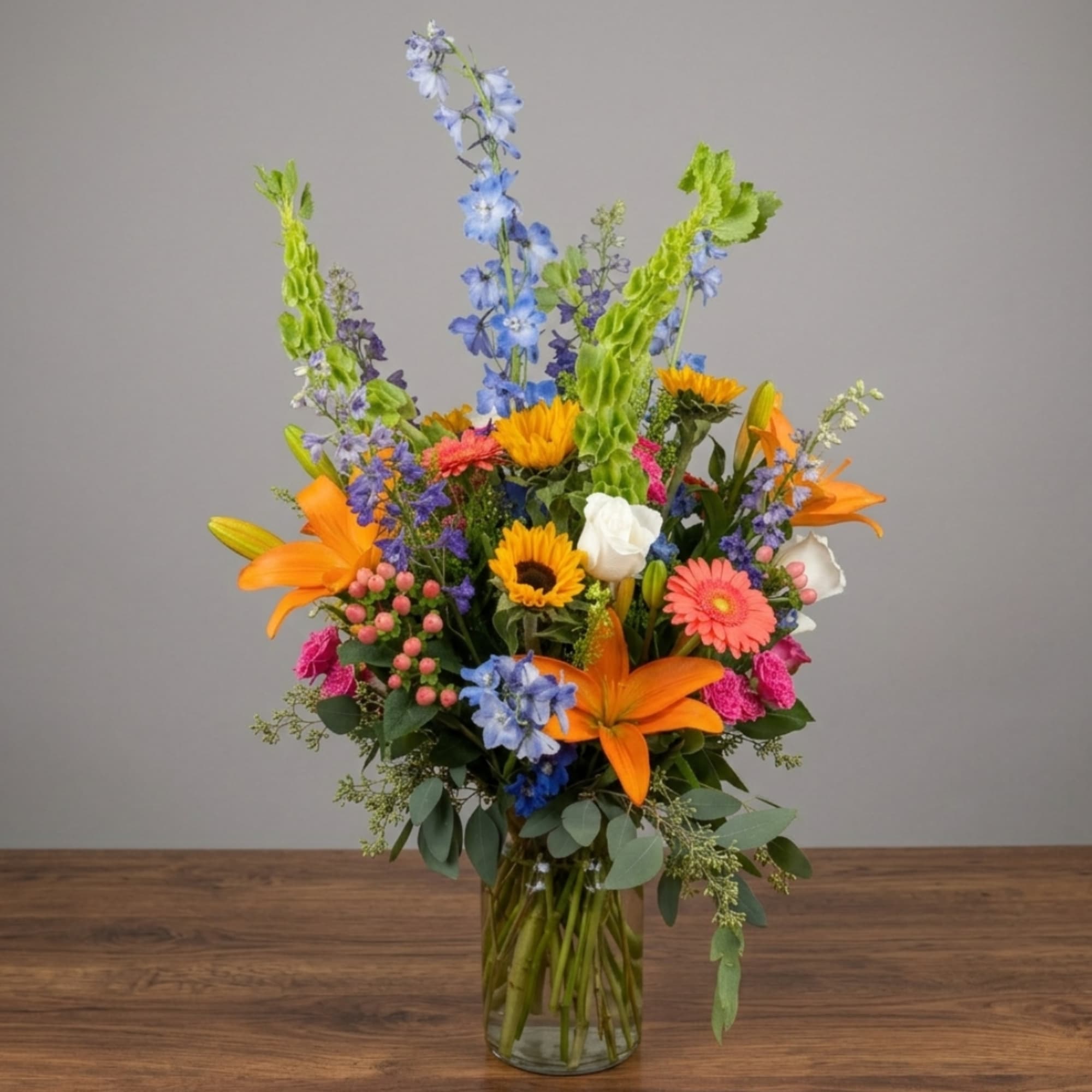 Mixed bouquet with orange lilies, blue delphinium, and sunflowers in a glass vase.
