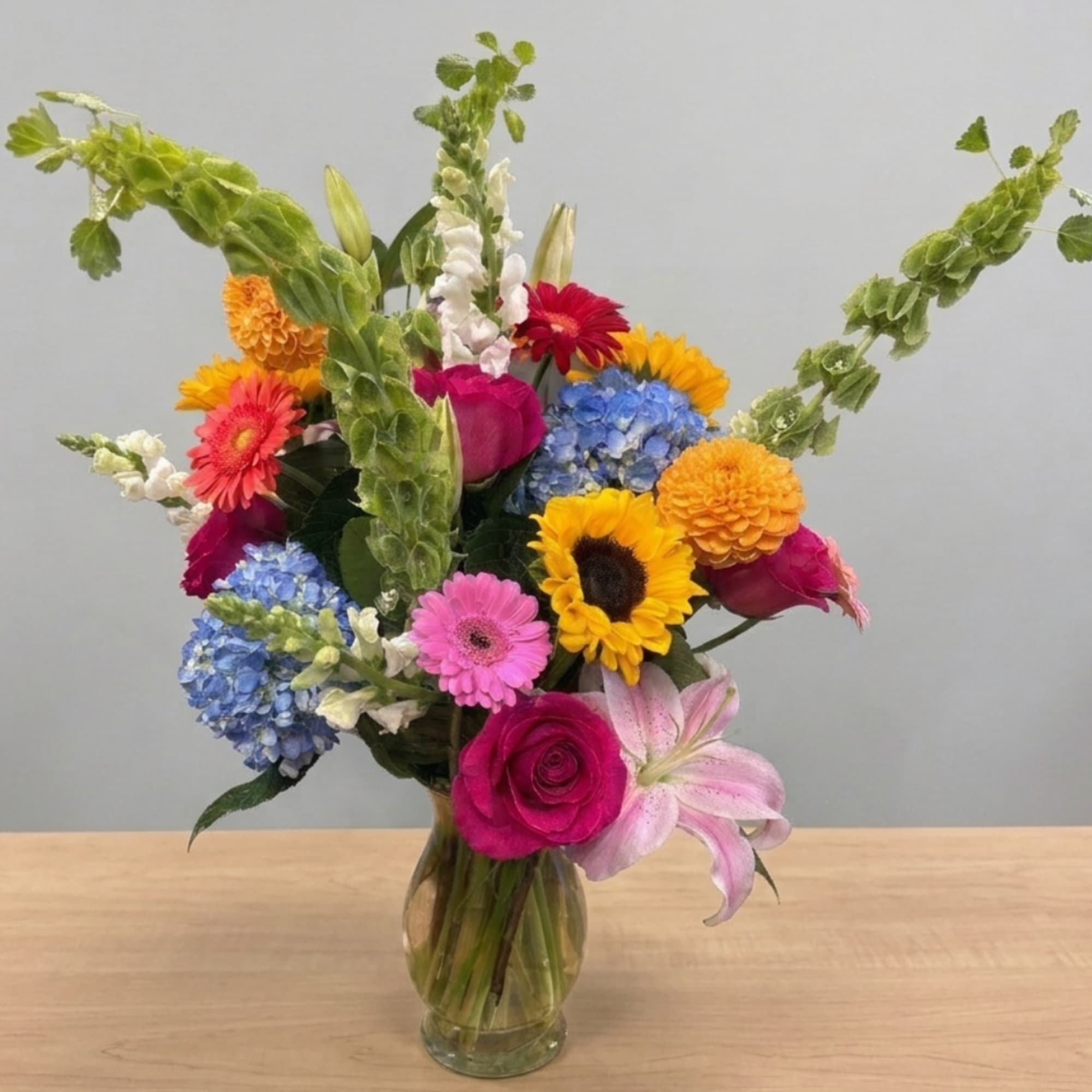 Colorful mixed bouquet in a clear glass vase with sunflowers, roses, lilies, and blue hydrangeas