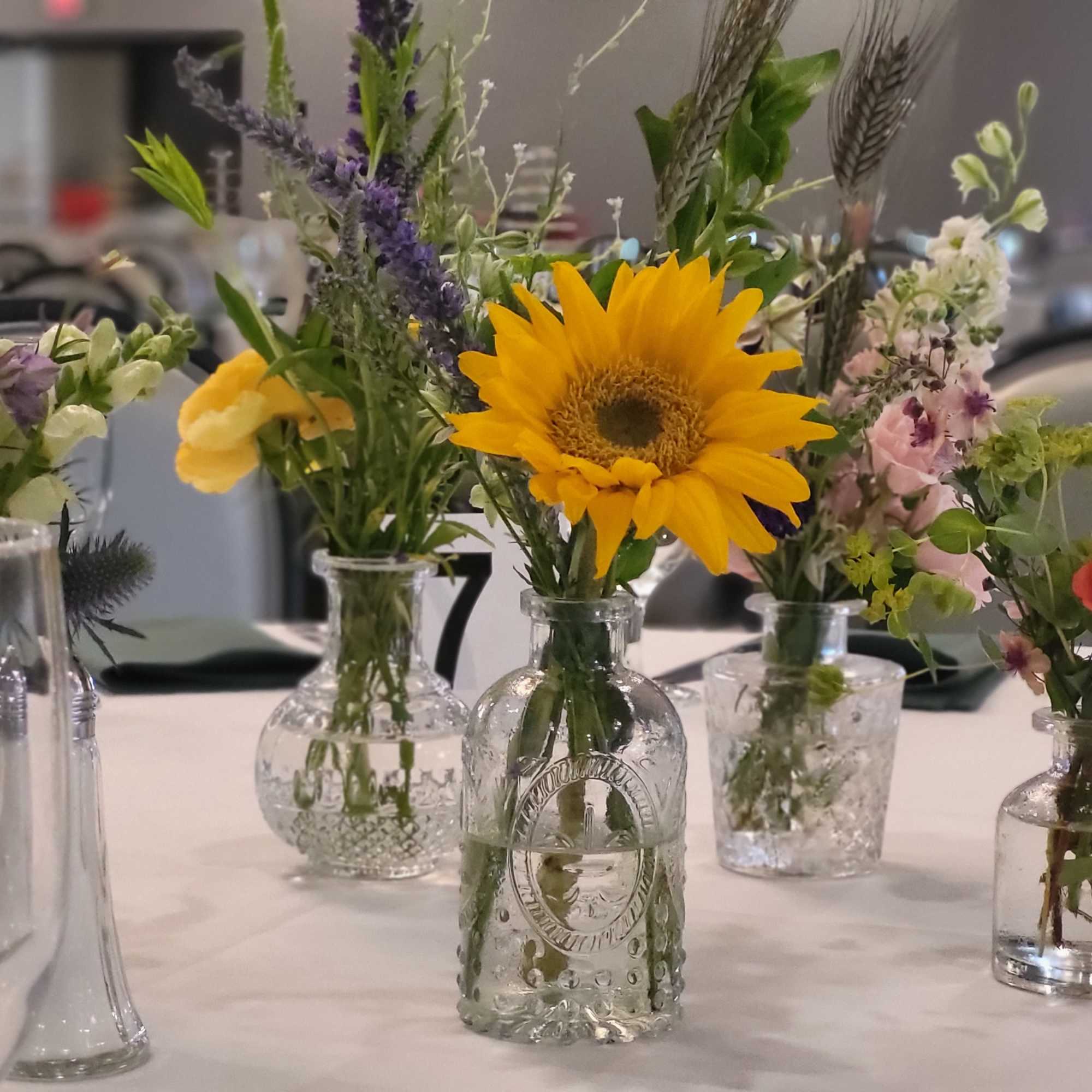 Small mixed flower arrangements in glass bottles on a table, with a sunflower centerpiece