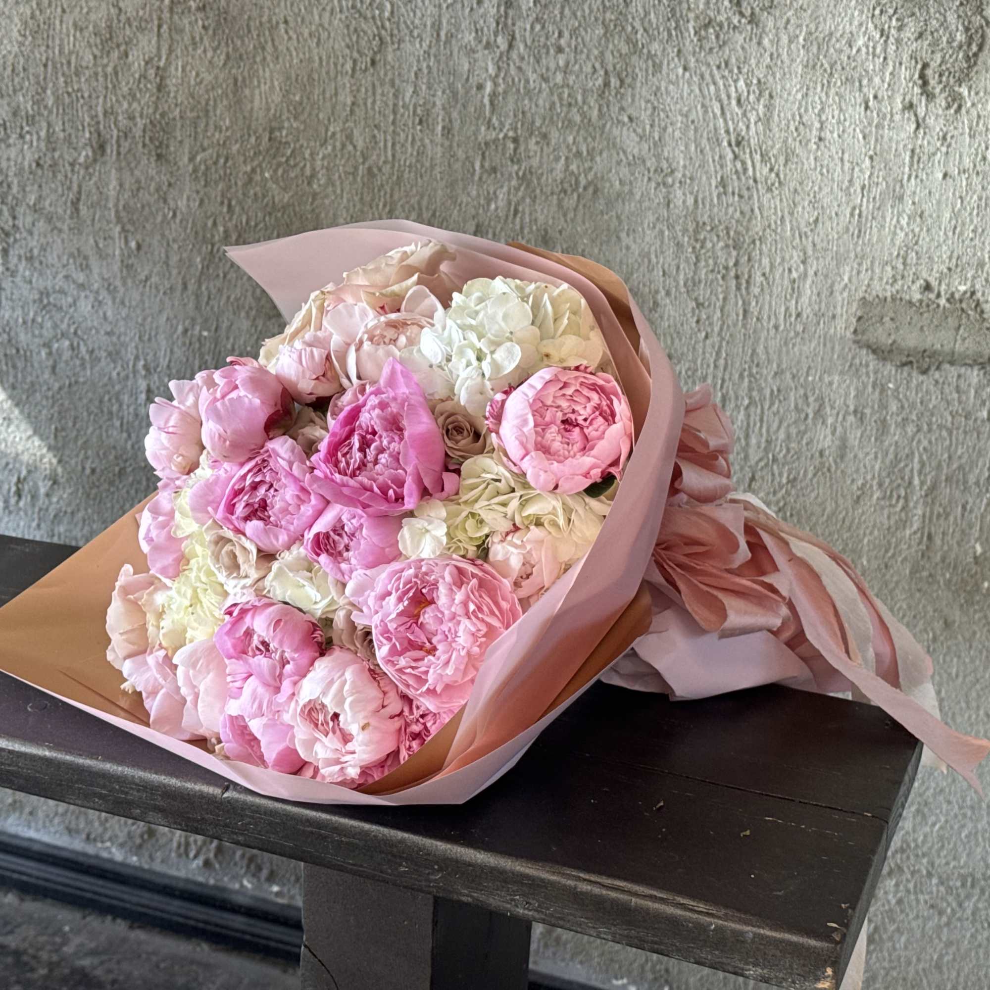 Hand-tied bouquet of pink and white peonies wrapped in pink and tan paper with ribbons.