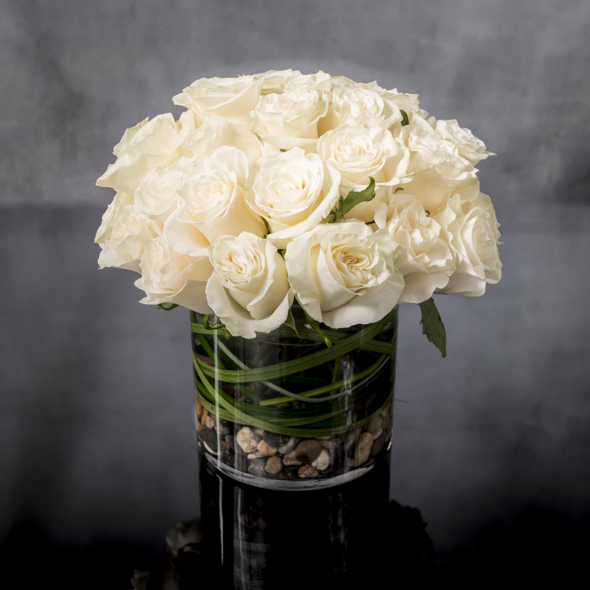 Compact arrangement of white roses in a clear glass cylinder vase with stones at the base