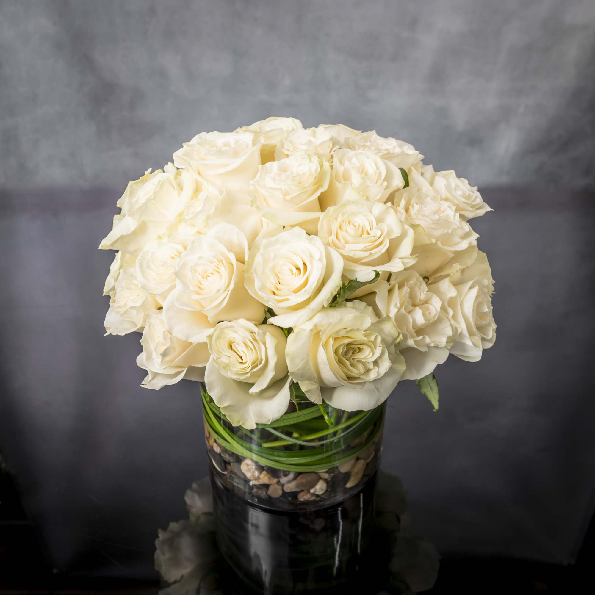 Compact arrangement of cream roses in a clear glass cylinder vase with pebbles at the base.
