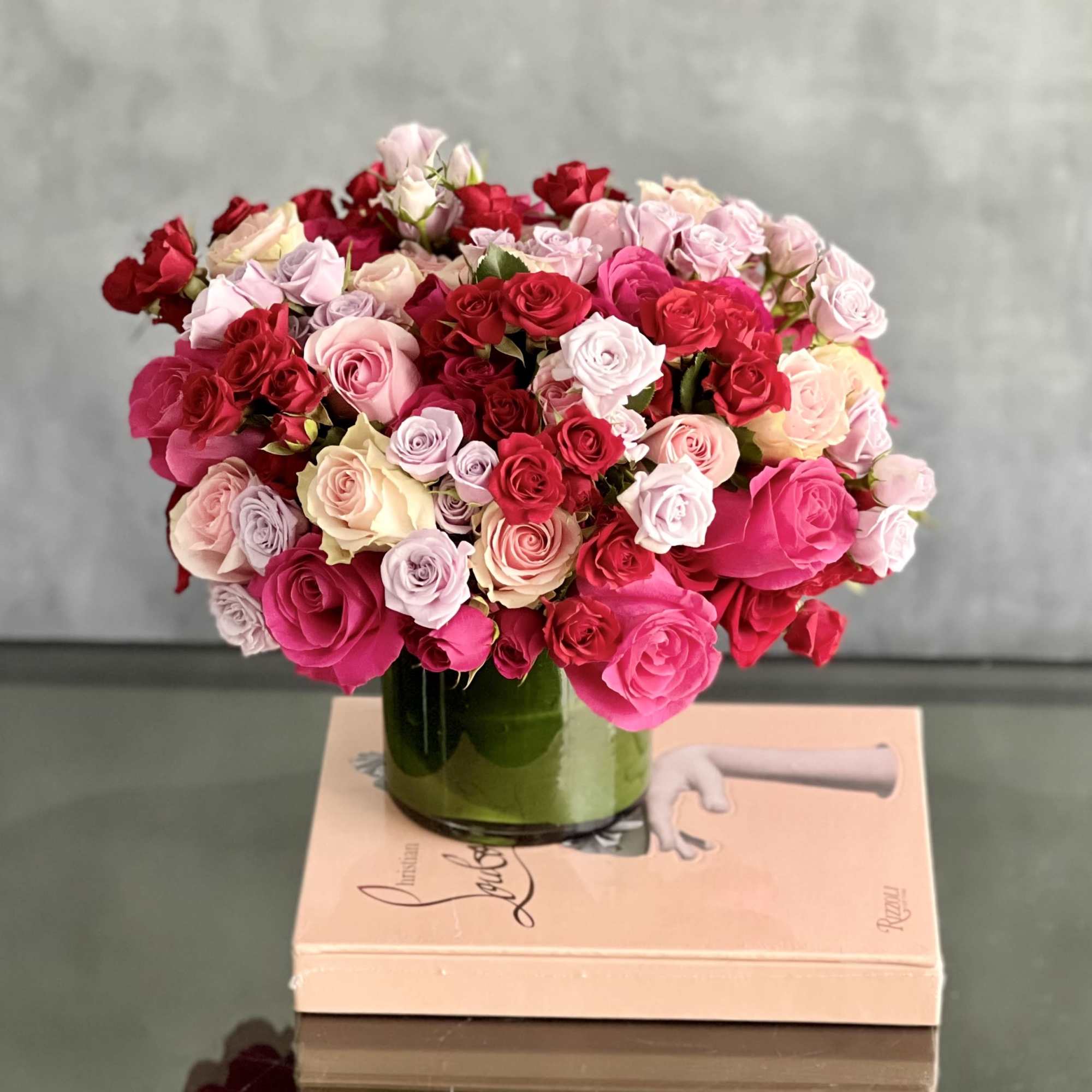 Low glass vase filled with dense red, pink, and pastel roses arranged on a book.