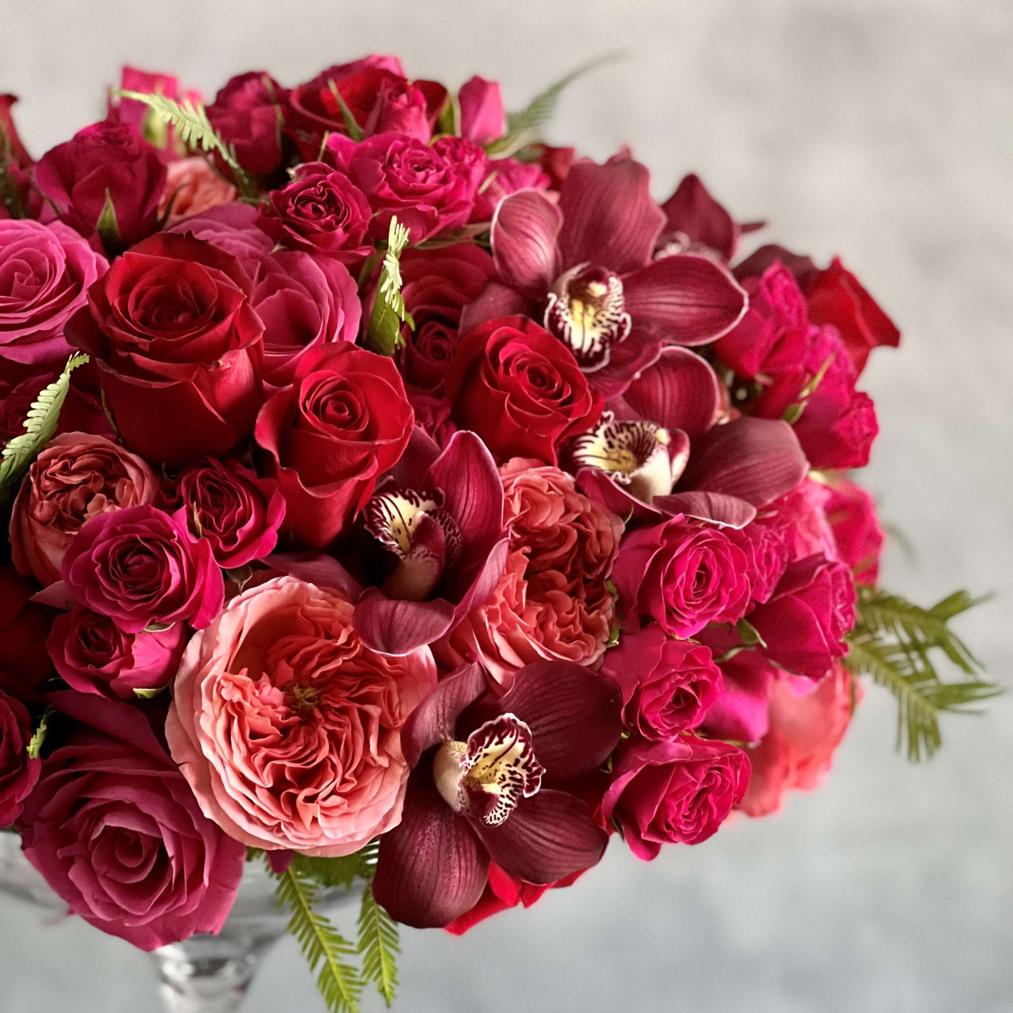 Dense arrangement of red and hot pink roses with burgundy orchids in a clear glass vase