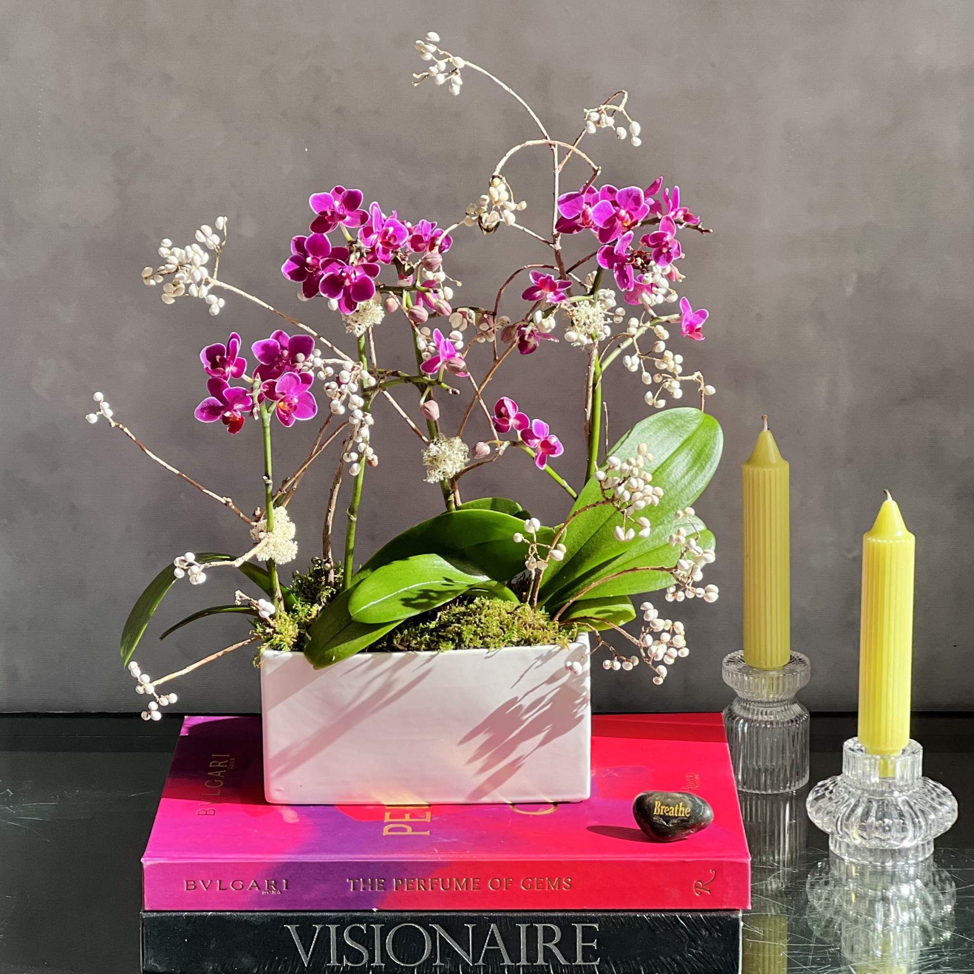 Pink orchid arrangement in a white rectangular planter beside two yellow candles, photo 3 of 4