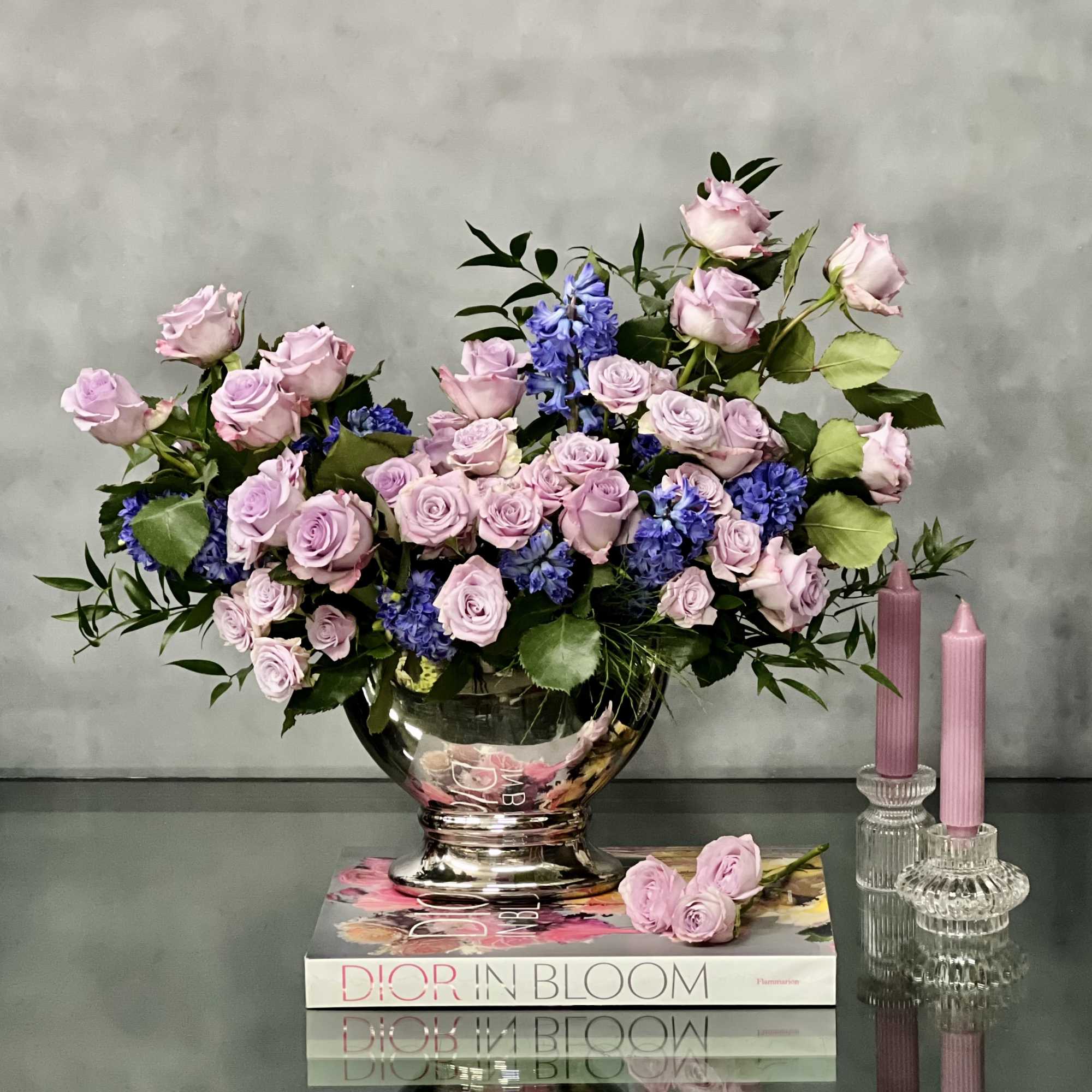 Lavender roses and blue flowers arranged in a silver bowl on books beside pink taper candles
