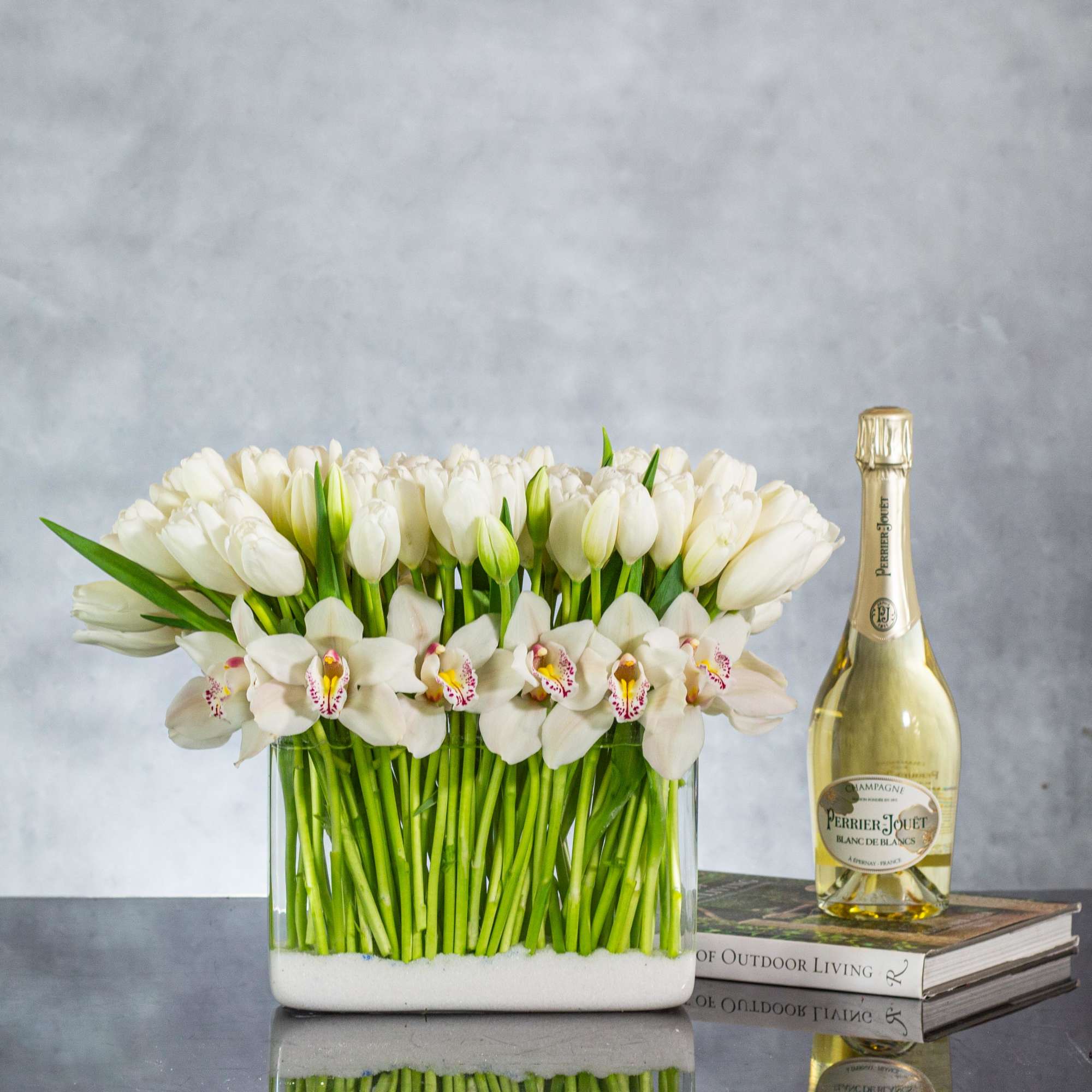 Rectangular vase of white tulips and orchids with a champagne bottle beside it on stacked books