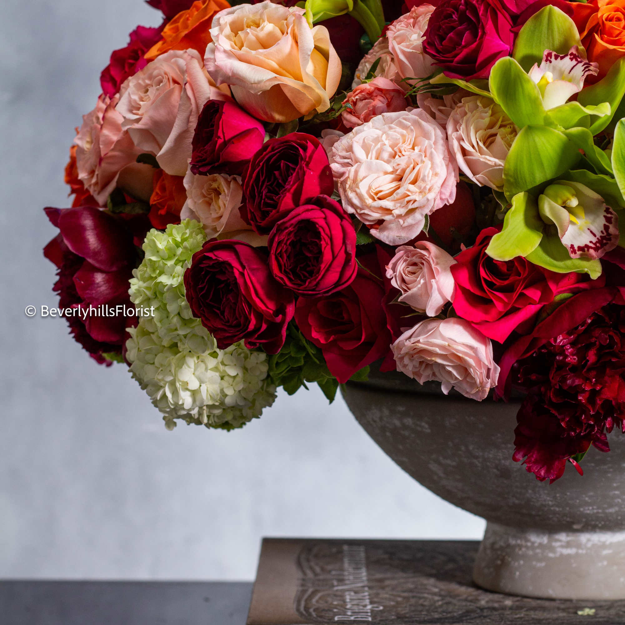 Mixed bouquet of red, pink, peach, and green flowers in a pedestal vase
