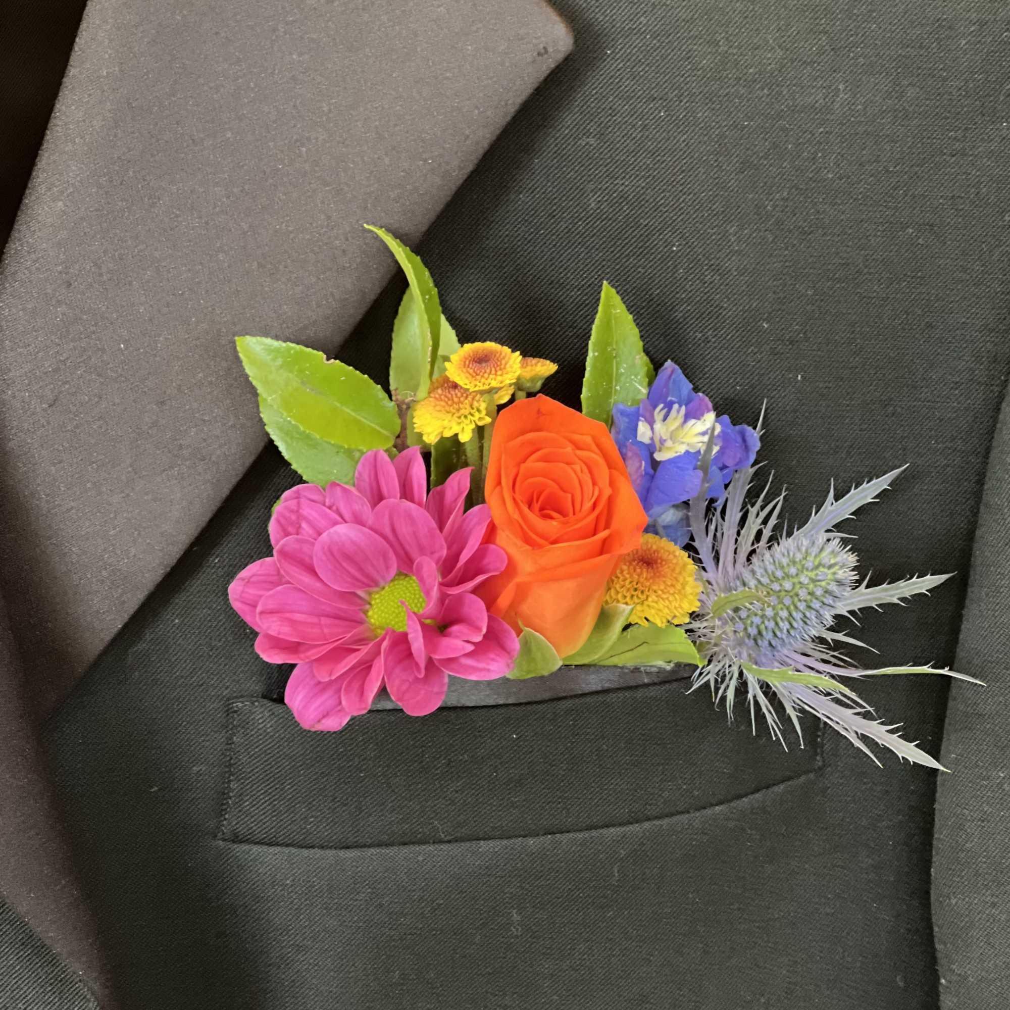 Colorful pocket boutonniere with orange rose, pink daisy, and blue thistle on a dark suit.