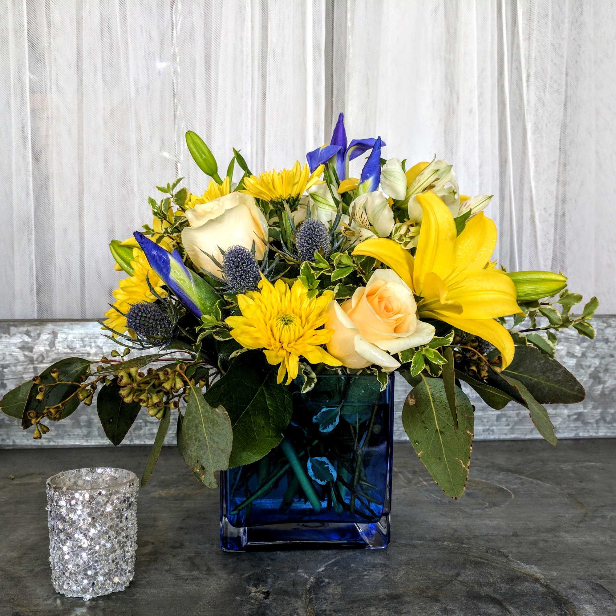 Compact arrangement of yellow lilies, cream roses, and blue accents in a blue glass cube vase.