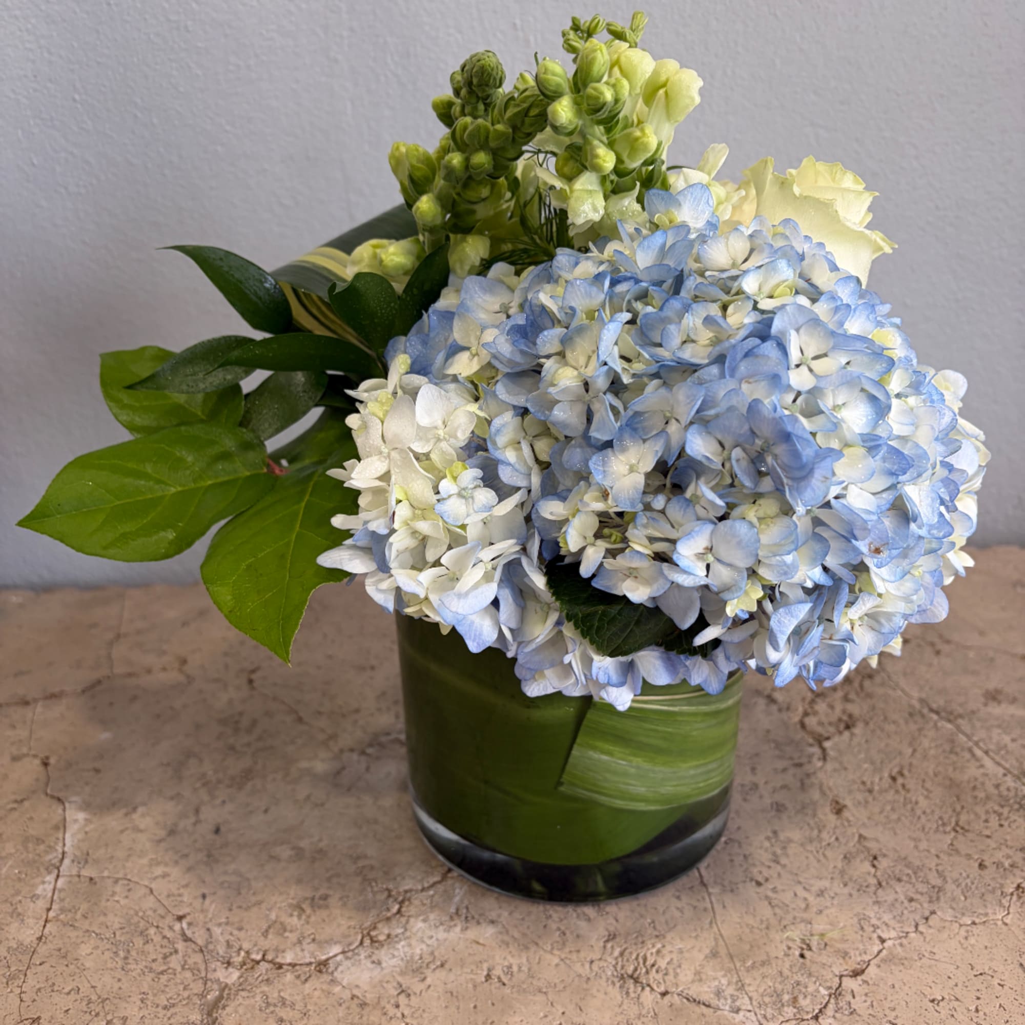 Blue and white hydrangeas in a green glass vase with broad leaves