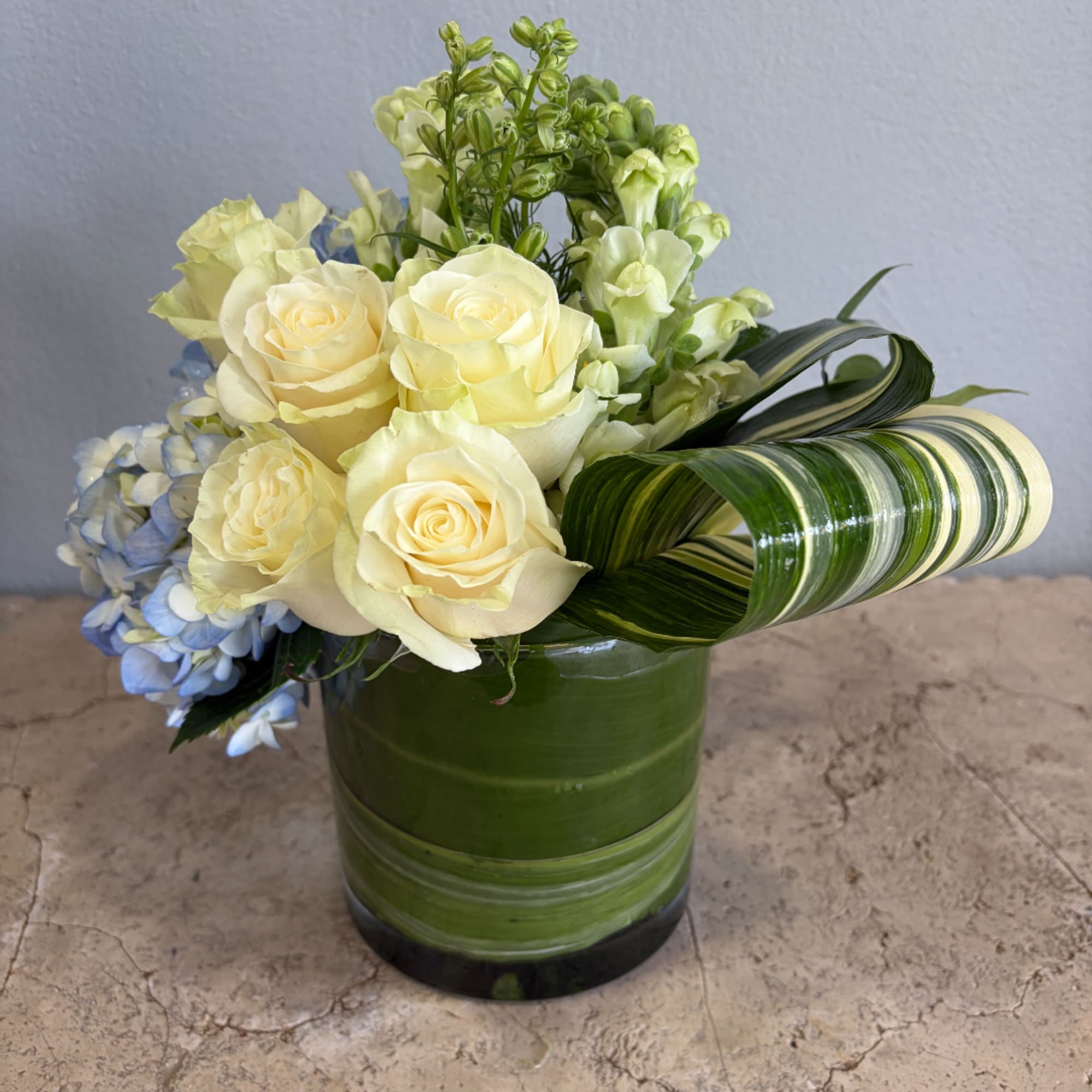 Cream roses and blue hydrangea in a green glass vase with striped leaves