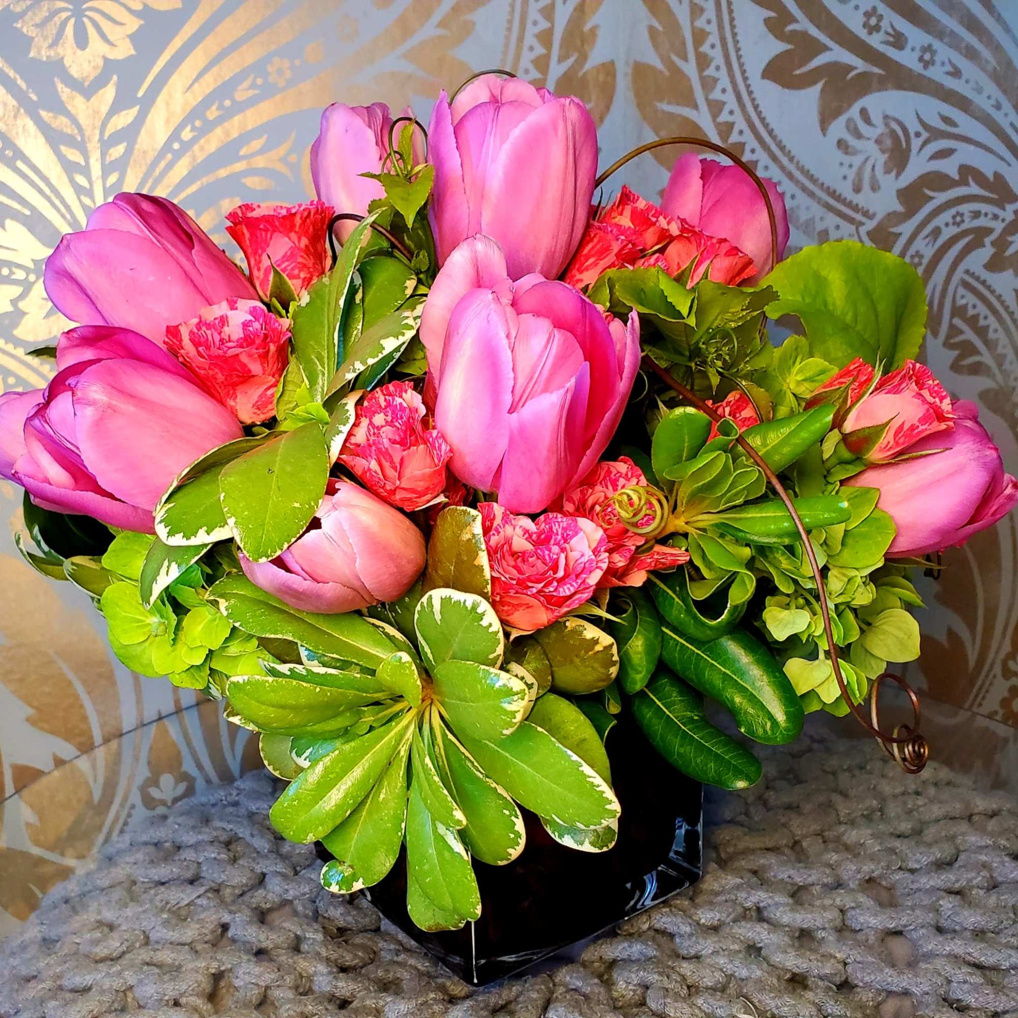 Compact arrangement of pink tulips and pink carnations with variegated green foliage in a black cube vase