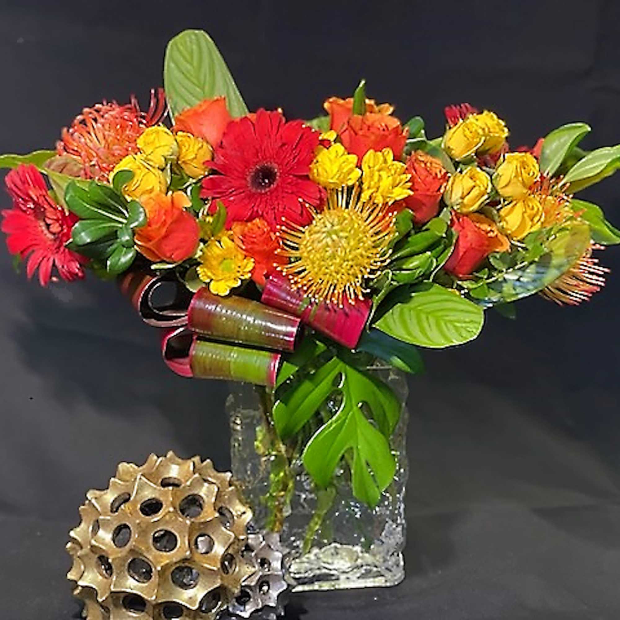 Bright arrangement of red gerbera daisies, roses and yellow pincushion blooms in a clear glass vase.