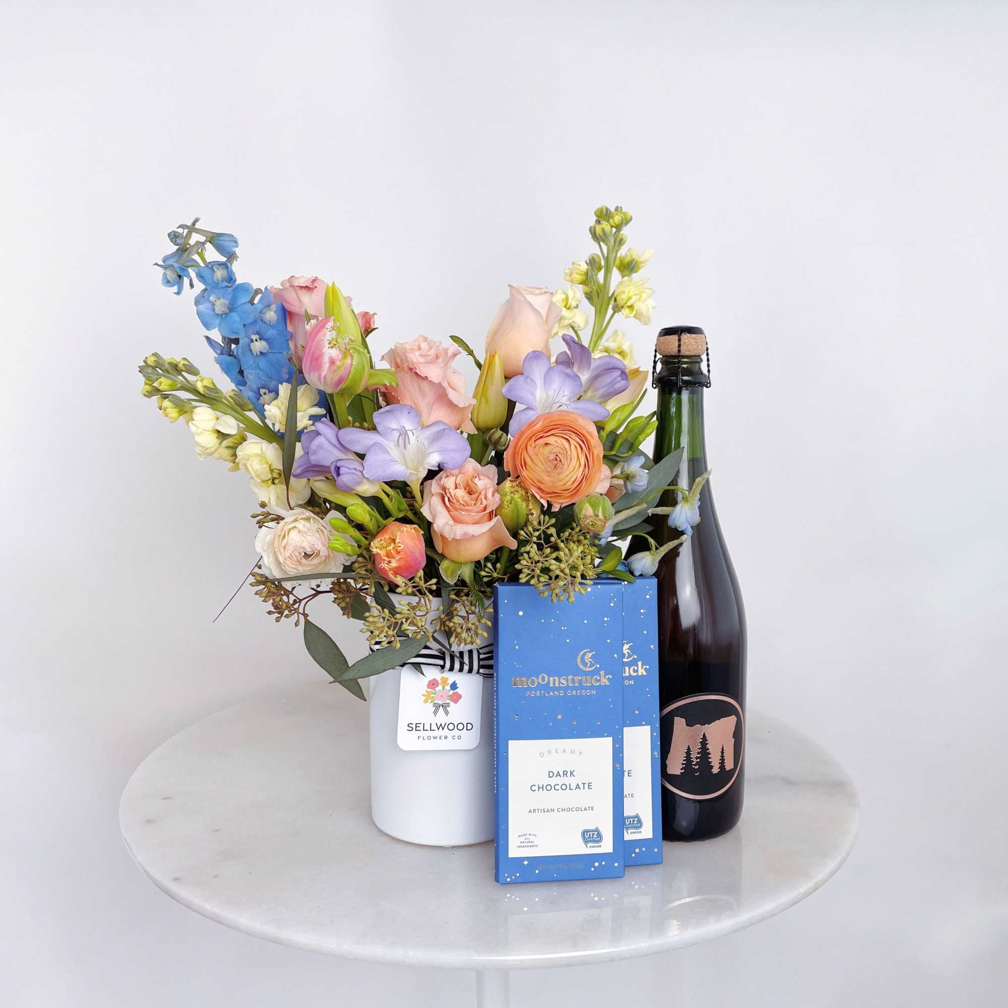 Mixed pastel bouquet in a white vase with chocolate boxes and a bottle of sparkling wine