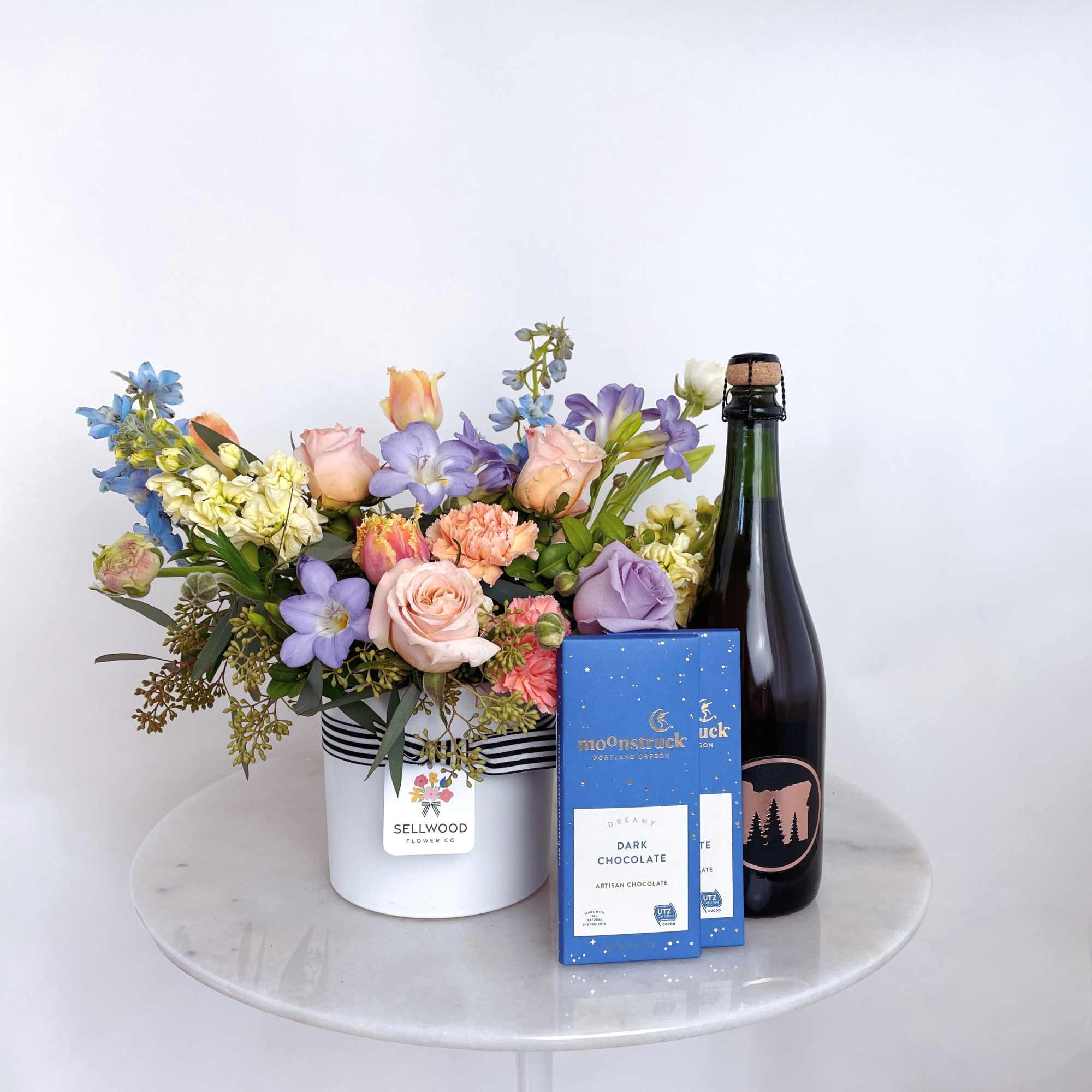 Mixed pastel flower arrangement with chocolate bars and a bottle of sparkling wine