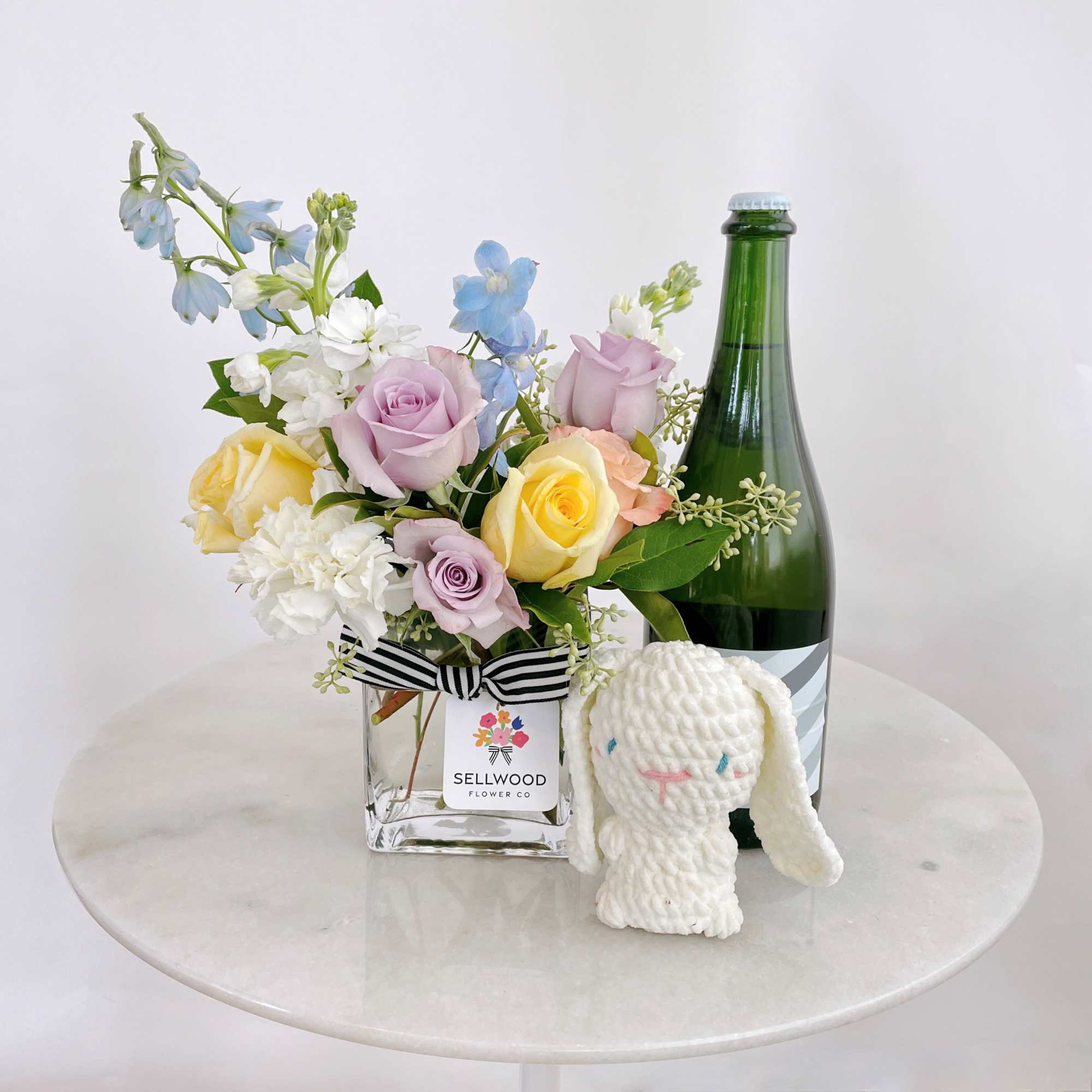 Mixed pastel flower arrangement in a square glass vase with a plush bunny and bottle