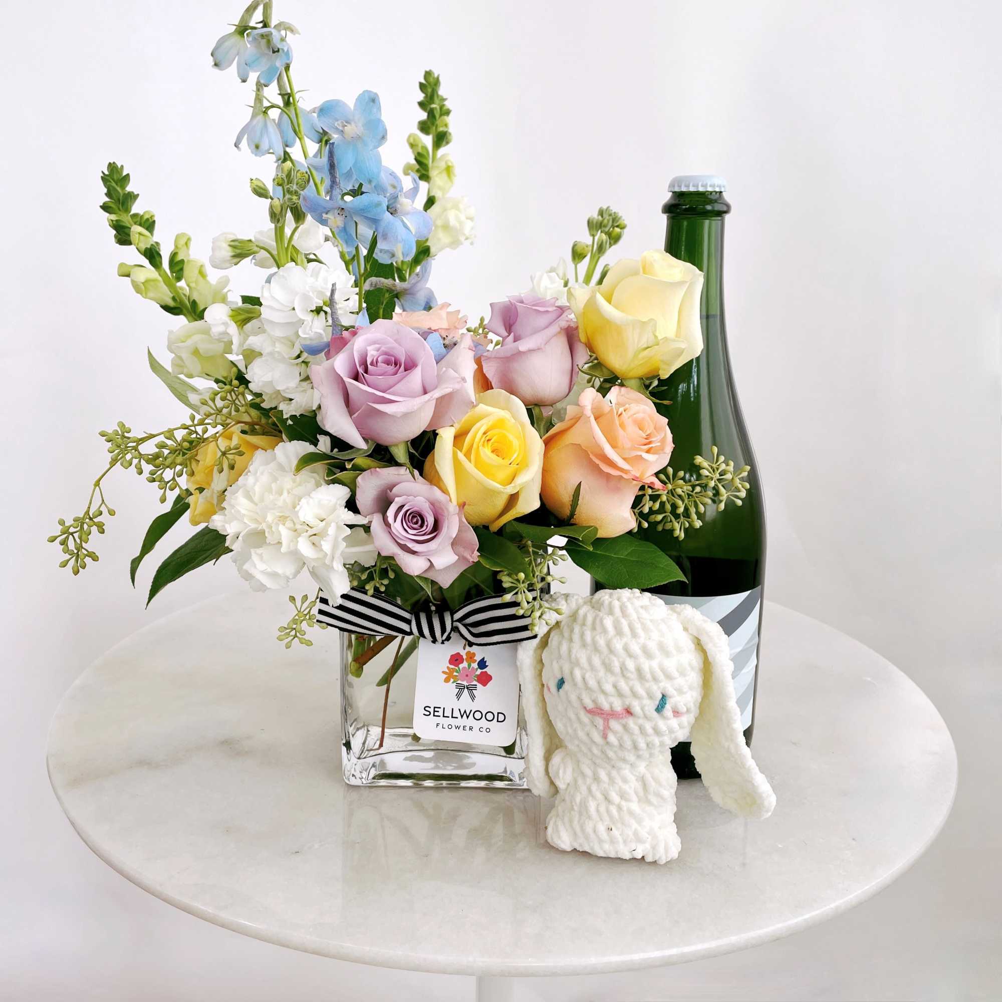 Pastel rose bouquet in a glass vase beside a green bottle and small plush bunny