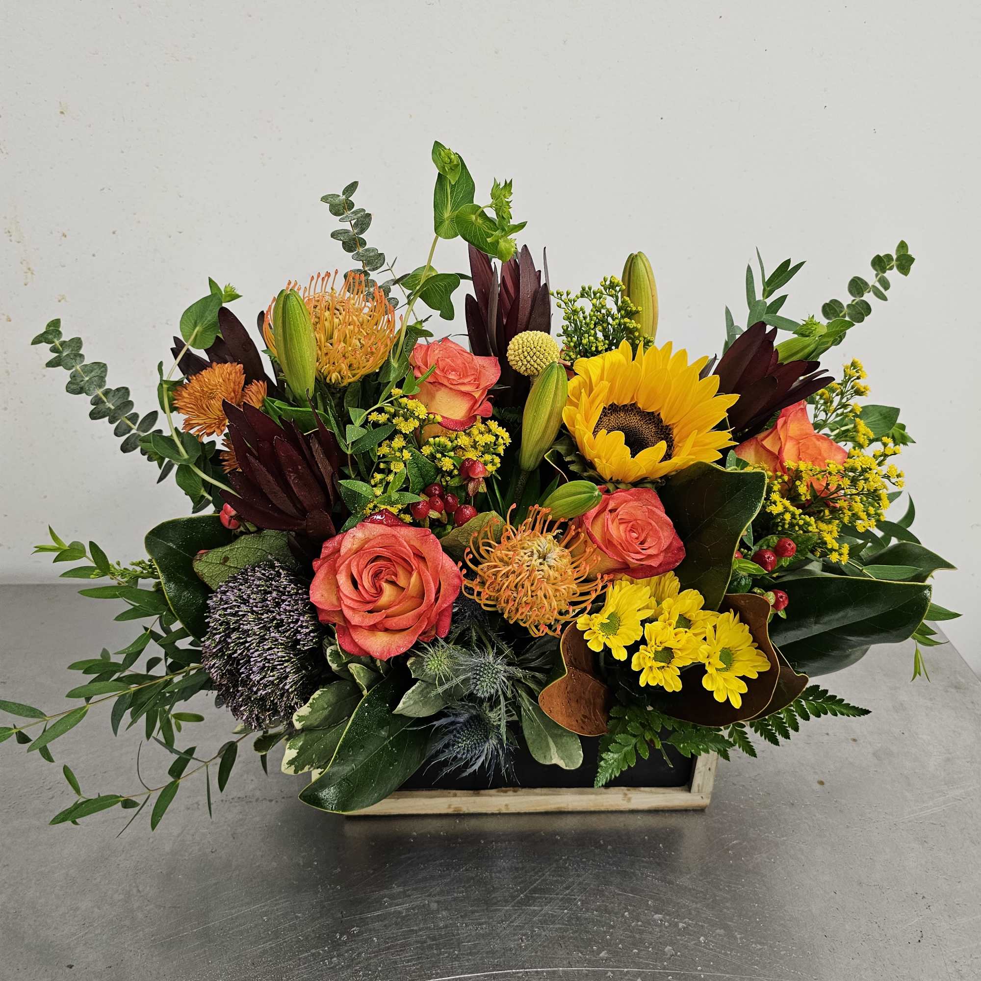 Mixed bouquet with roses, sunflowers, and orange blooms in a low wooden container